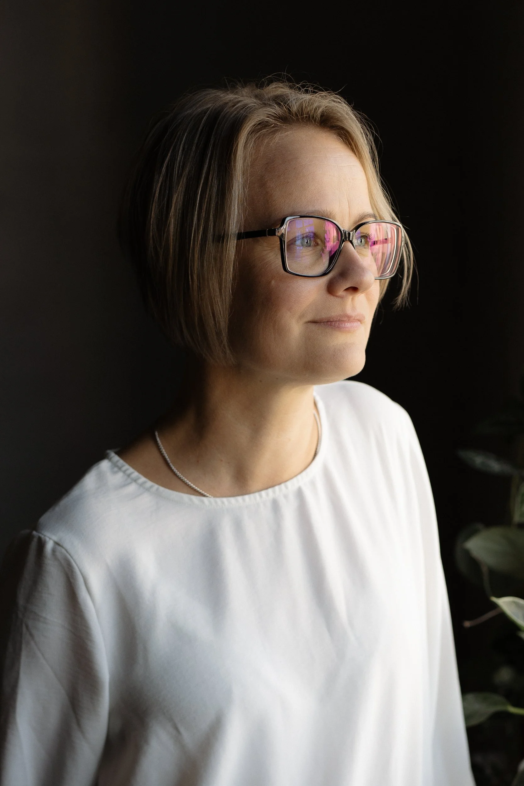 Lähikuva silmälaseja käyttävästä naisesta valkoisessa paidassa tummalla taustalla.