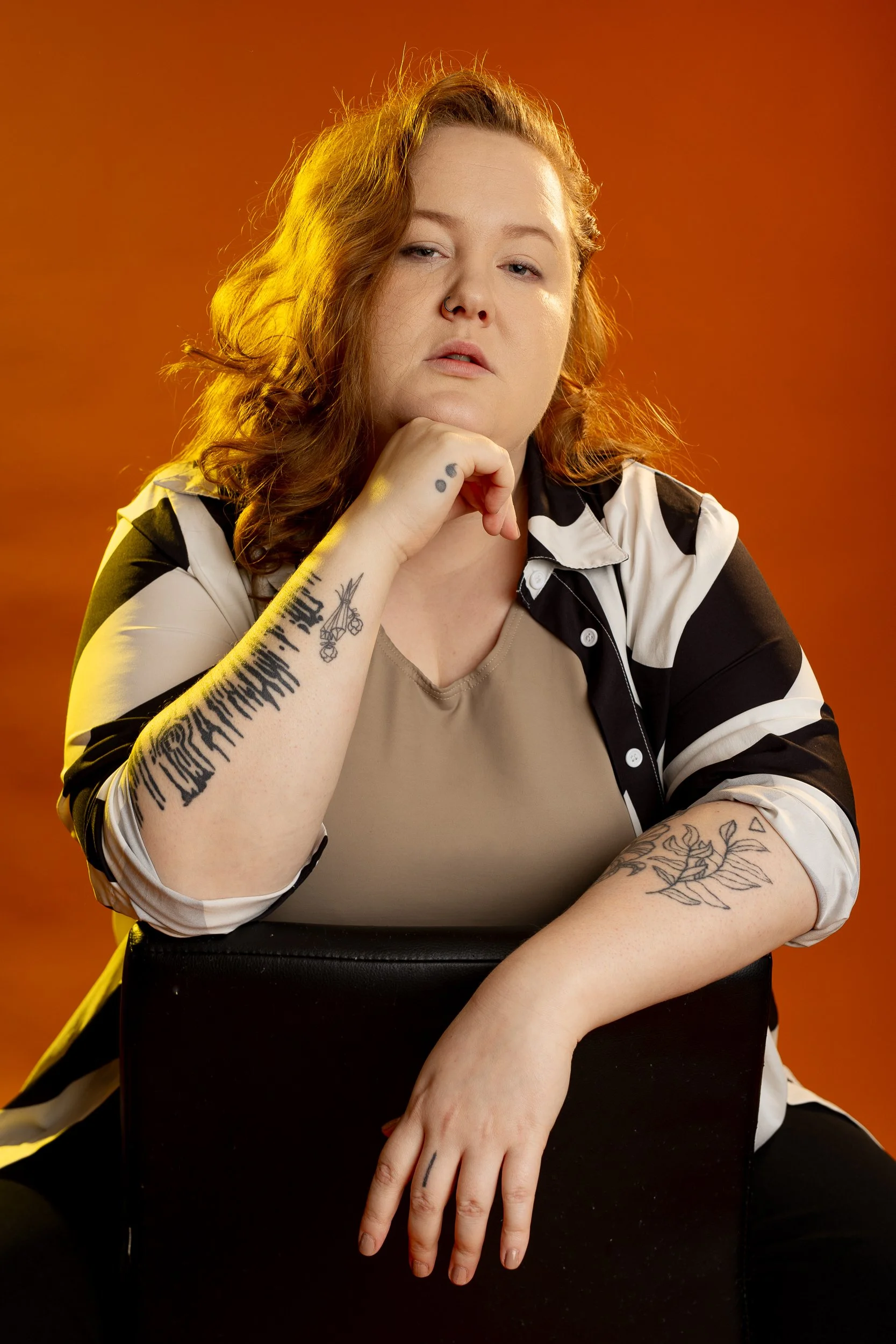Muotokuvassa tatuoitu nainen istuu mustalla tuolilla oranssia taustaa vasten.