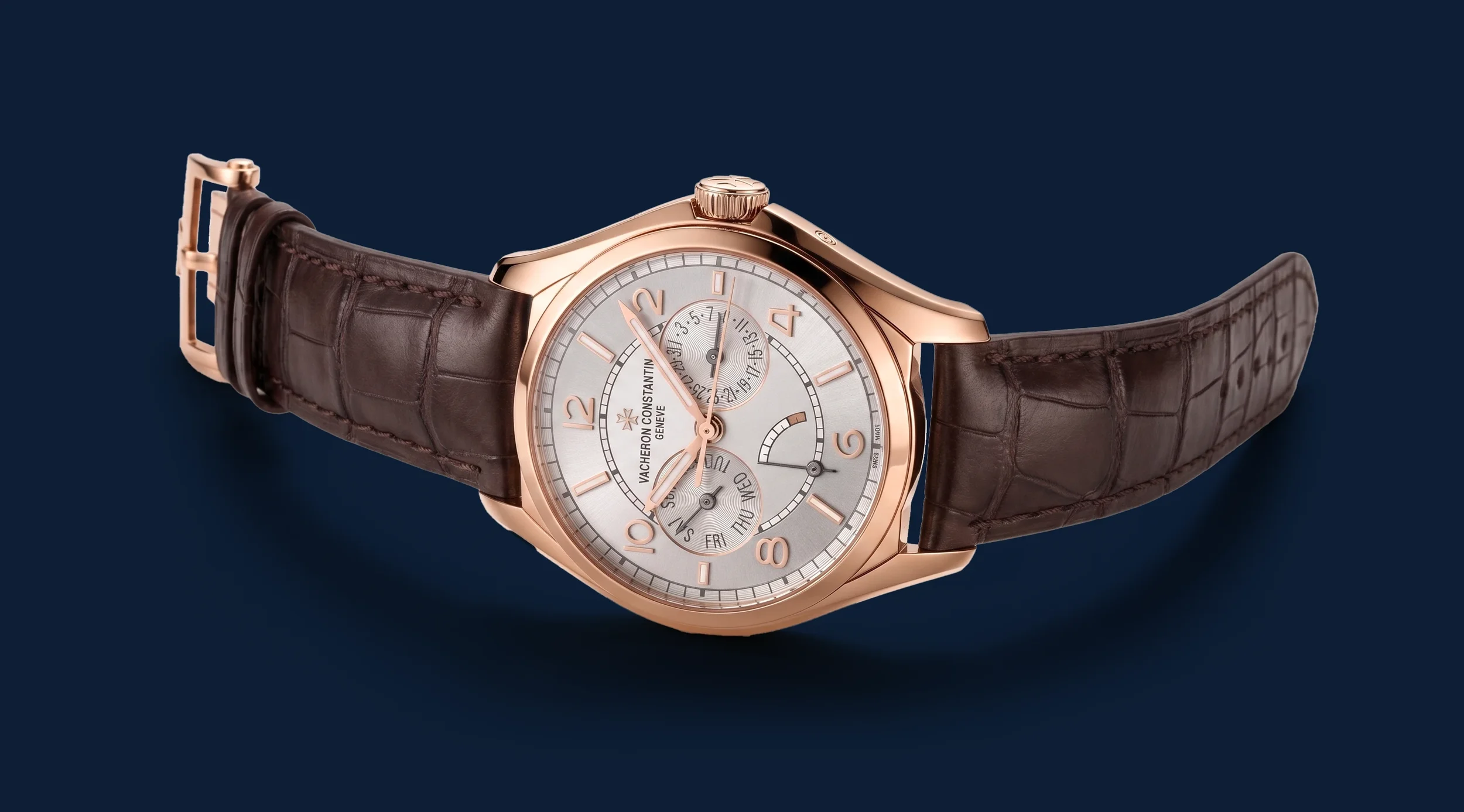 Close-up of a luxury wristwatch with a rose gold case, white dial, gold numerals, and a brown leather strap against a dark blue background.