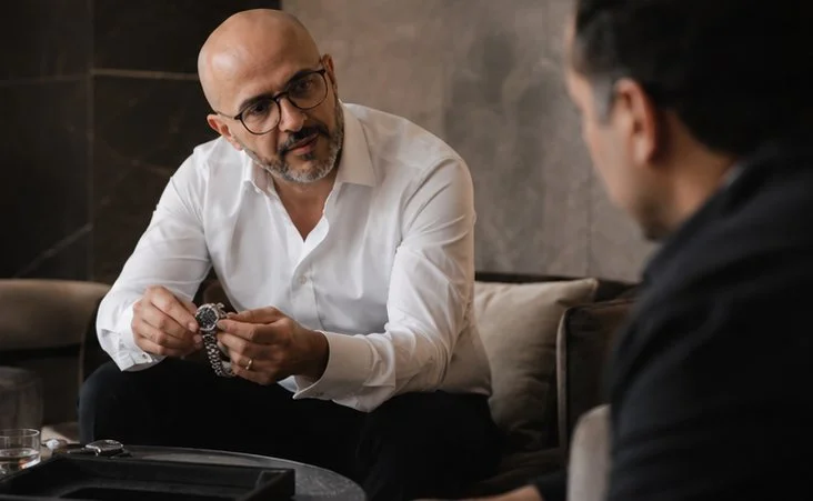 A bald man with glasses and a beard, wearing a white shirt, sitting on a sofa and holding a wristwatch, talking to another man with dark hair, wearing a dark shirt, seated across from him in a dimly lit room.
