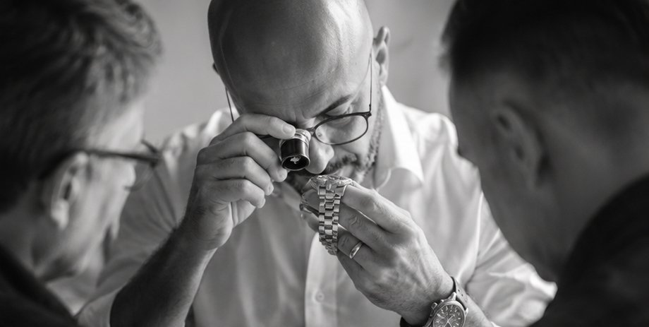 Three men closely examining a watch, with one using a magnifying tool and the others observing.