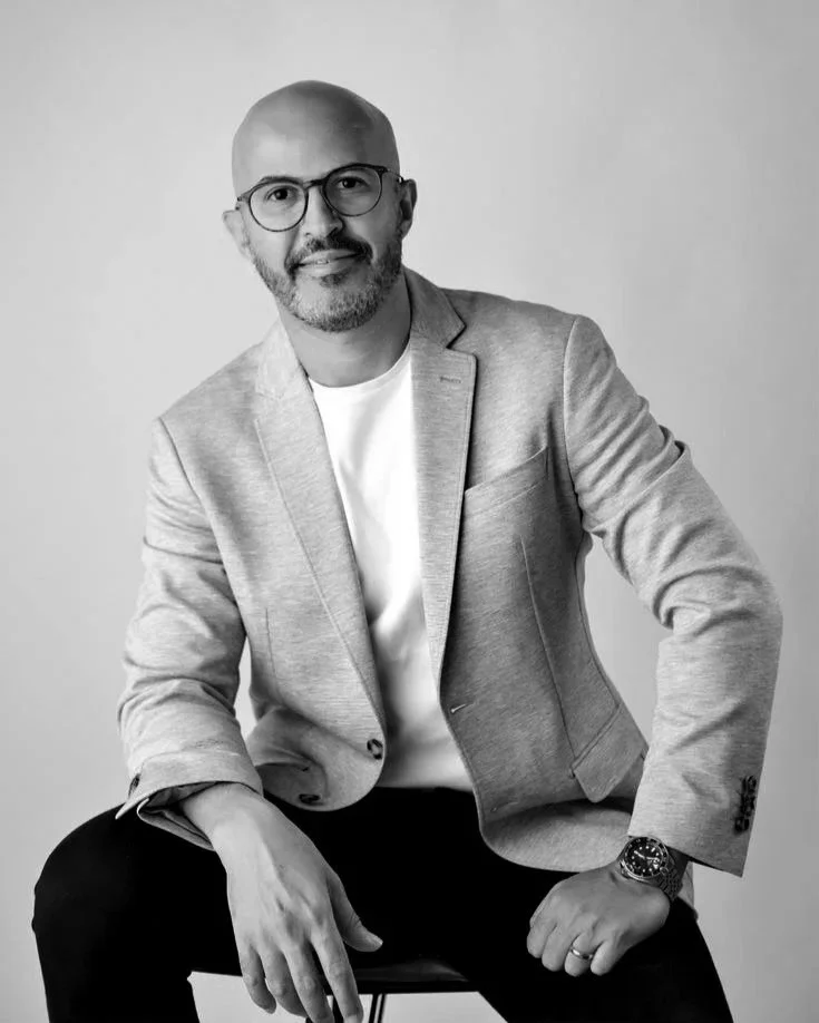 Man with glasses, short beard, wearing a light blazer over a white shirt, sitting on a stool against a plain background.