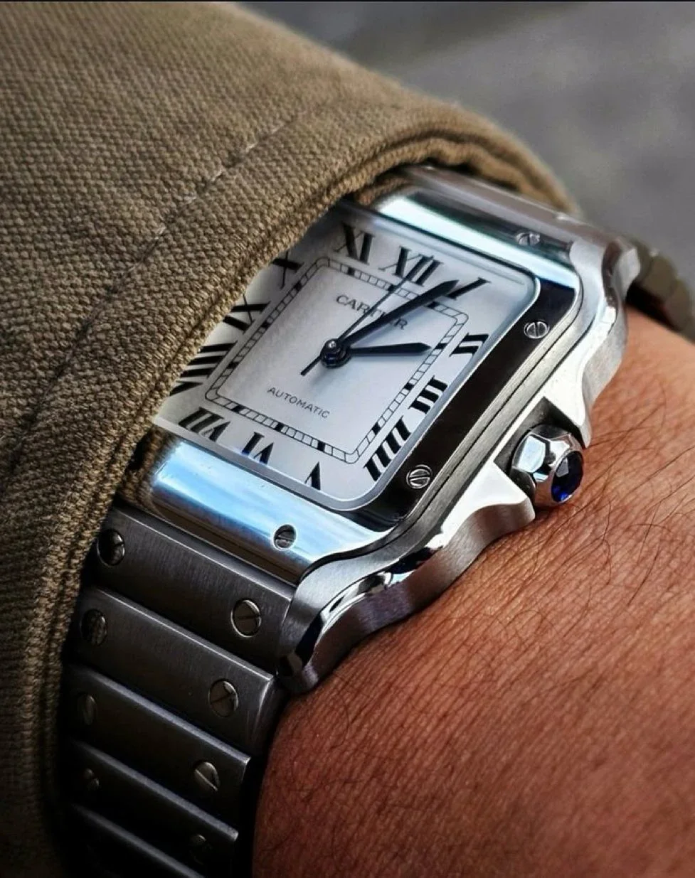 A close-up of a wrist wearing a luxury Cartier Tank watch with a square face, Roman numerals, and a stainless steel band, partially covered by a beige fabric sleeve.