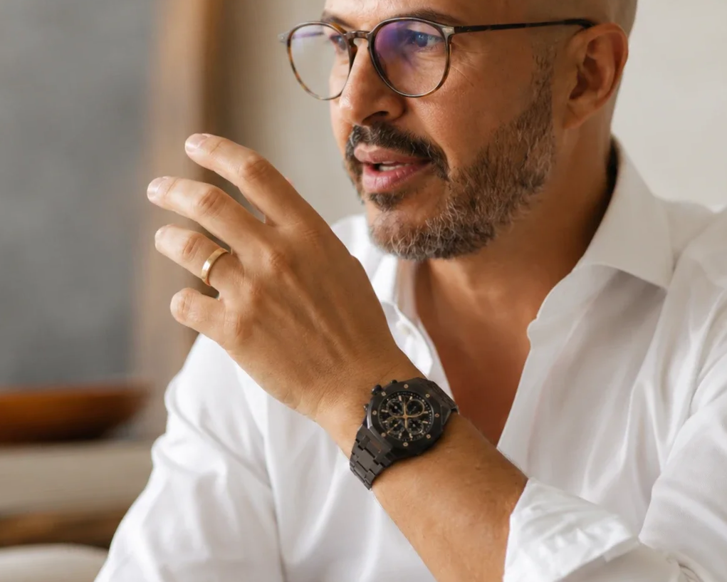 A middle-aged man with glasses and a beard, wearing a white shirt, is speaking or explaining something. He has a black wristwatch on his left wrist and a gold ring on his left hand. His hand is raised near his face, with fingers slightly spread.