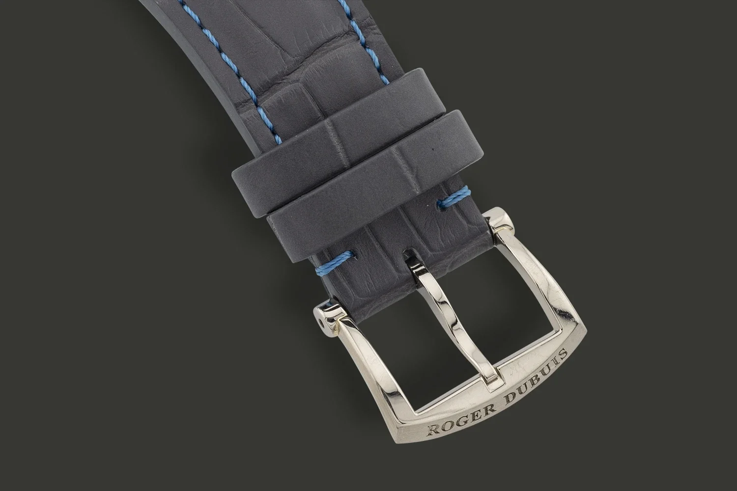 Close-up of a high-end watch strap with a silver buckle engraved with "ROGER DUBUIS," made of textured leather with blue stitching.