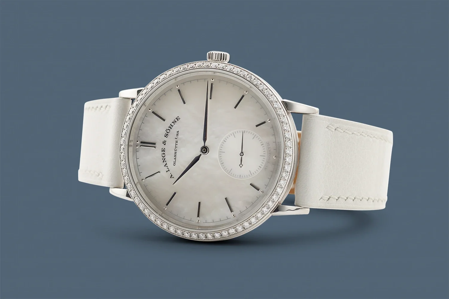 A luxury wristwatch with a white band, diamond-studded bezel, and a mother-of-pearl dial showing the time and a small seconds subdial, against a blue background.