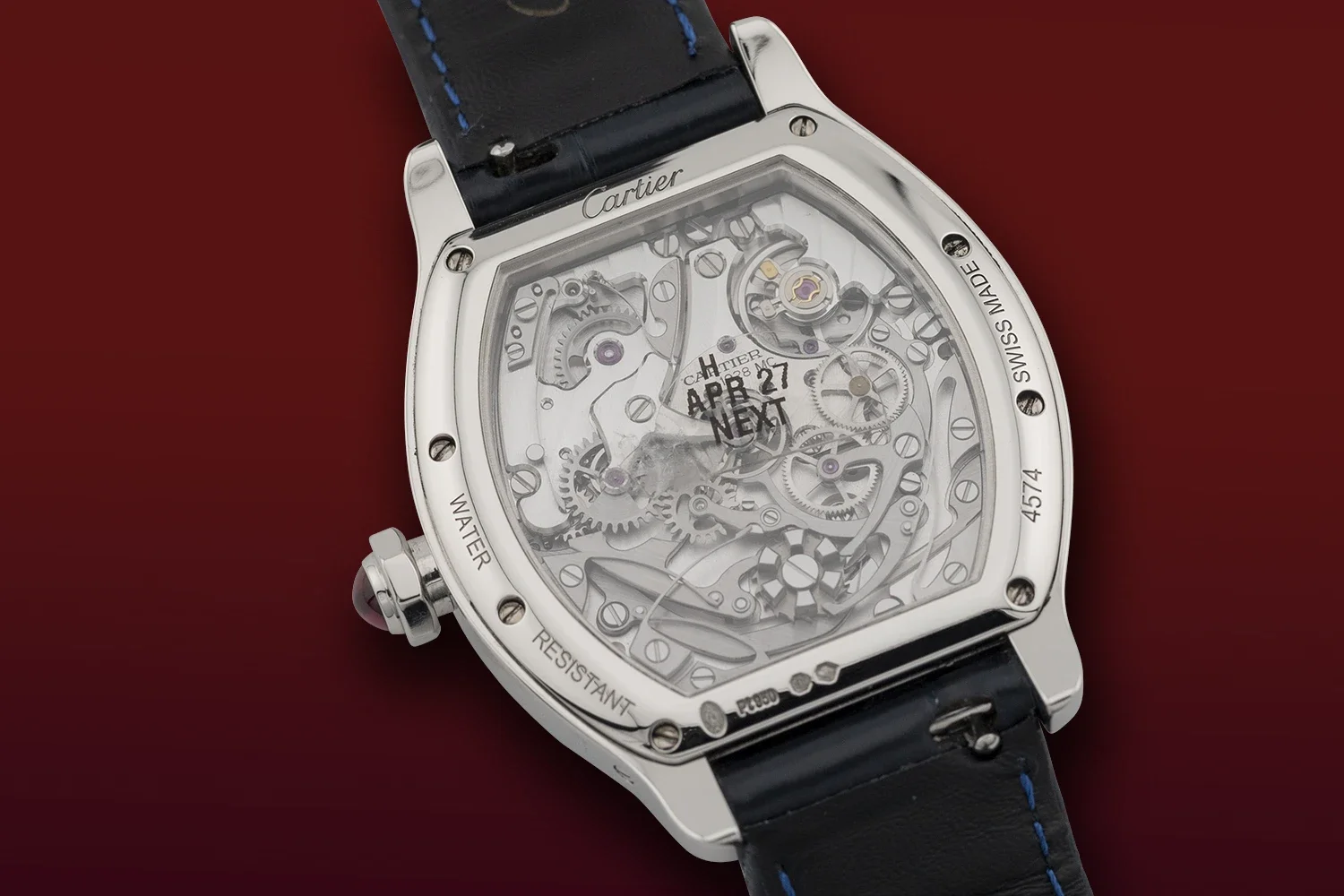 Close-up of a luxury Cartier skeleton watch with a visible mechanical movement, black leather strap with blue stitching, against a red background.