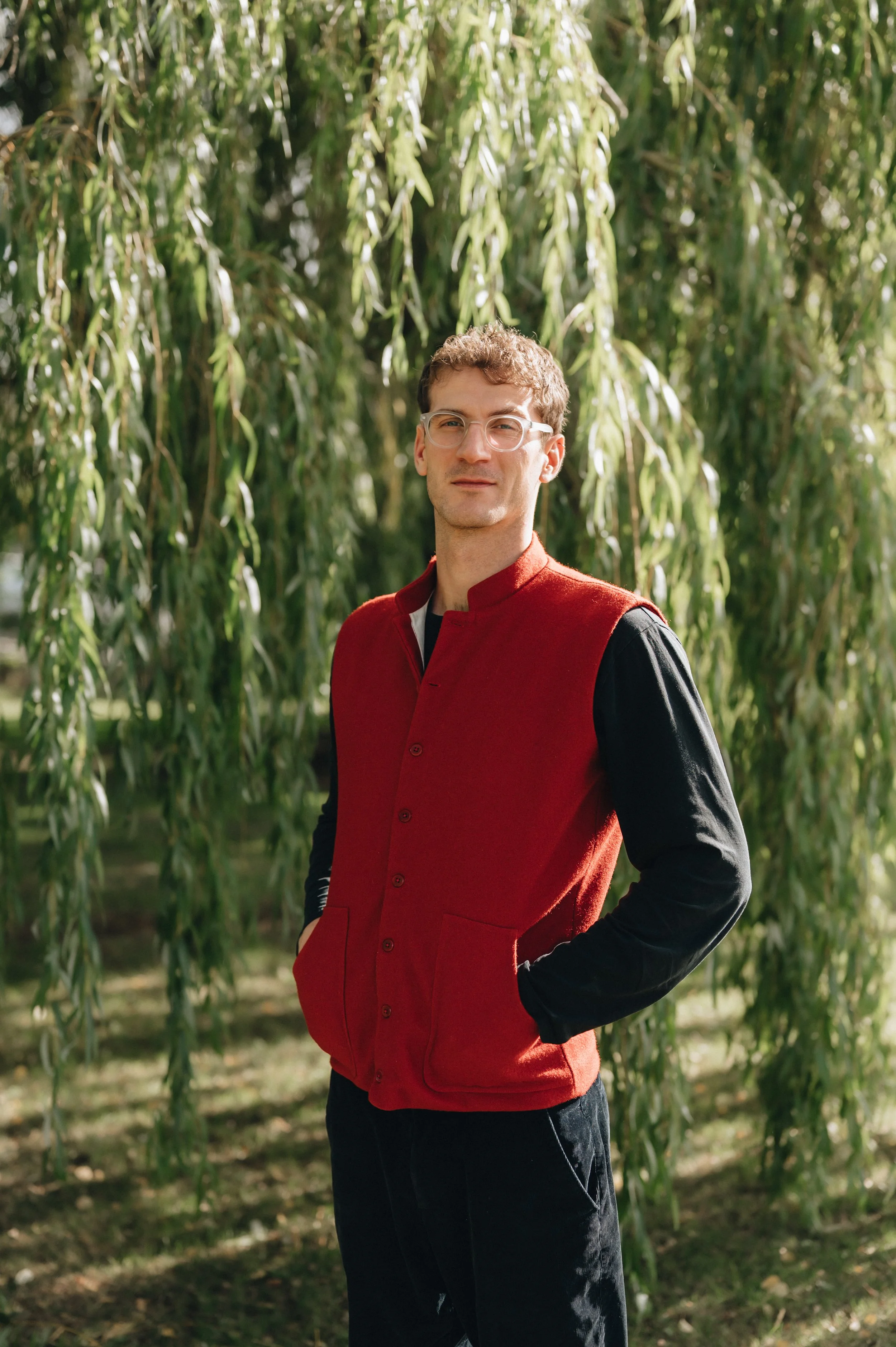 Man wearing white glasses and a red vest standing outdoors among tall green plants