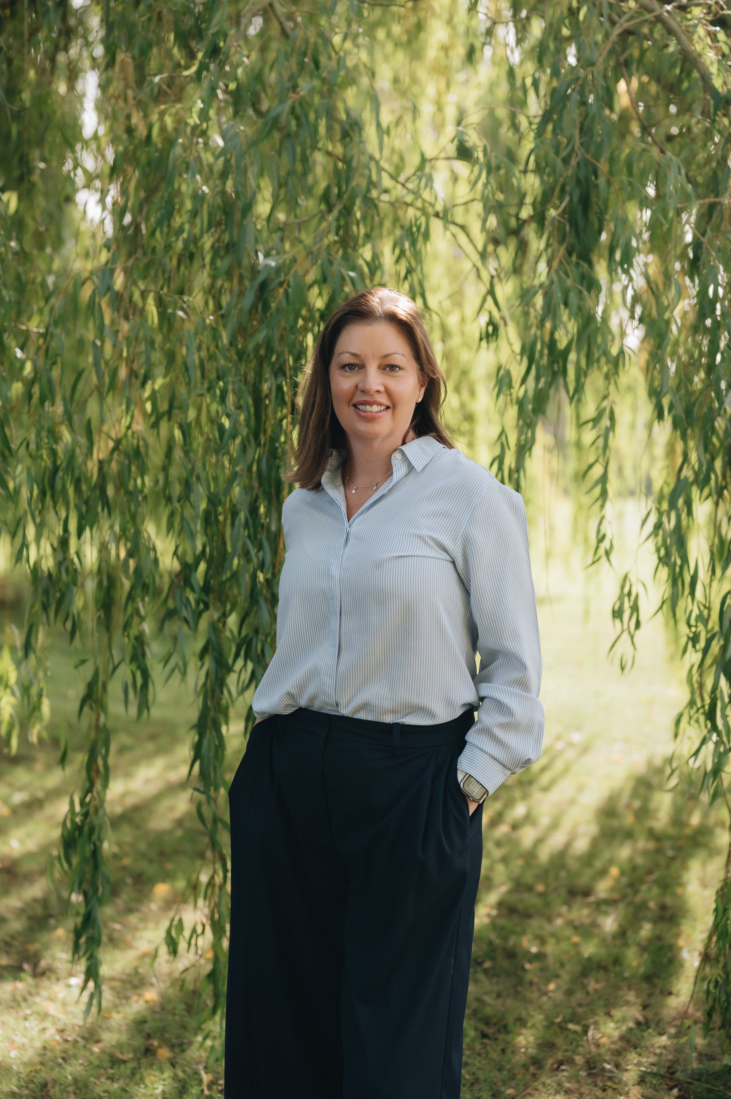 Smiling woman in a blue striped blouse standing outdoors among sunlit willow trees