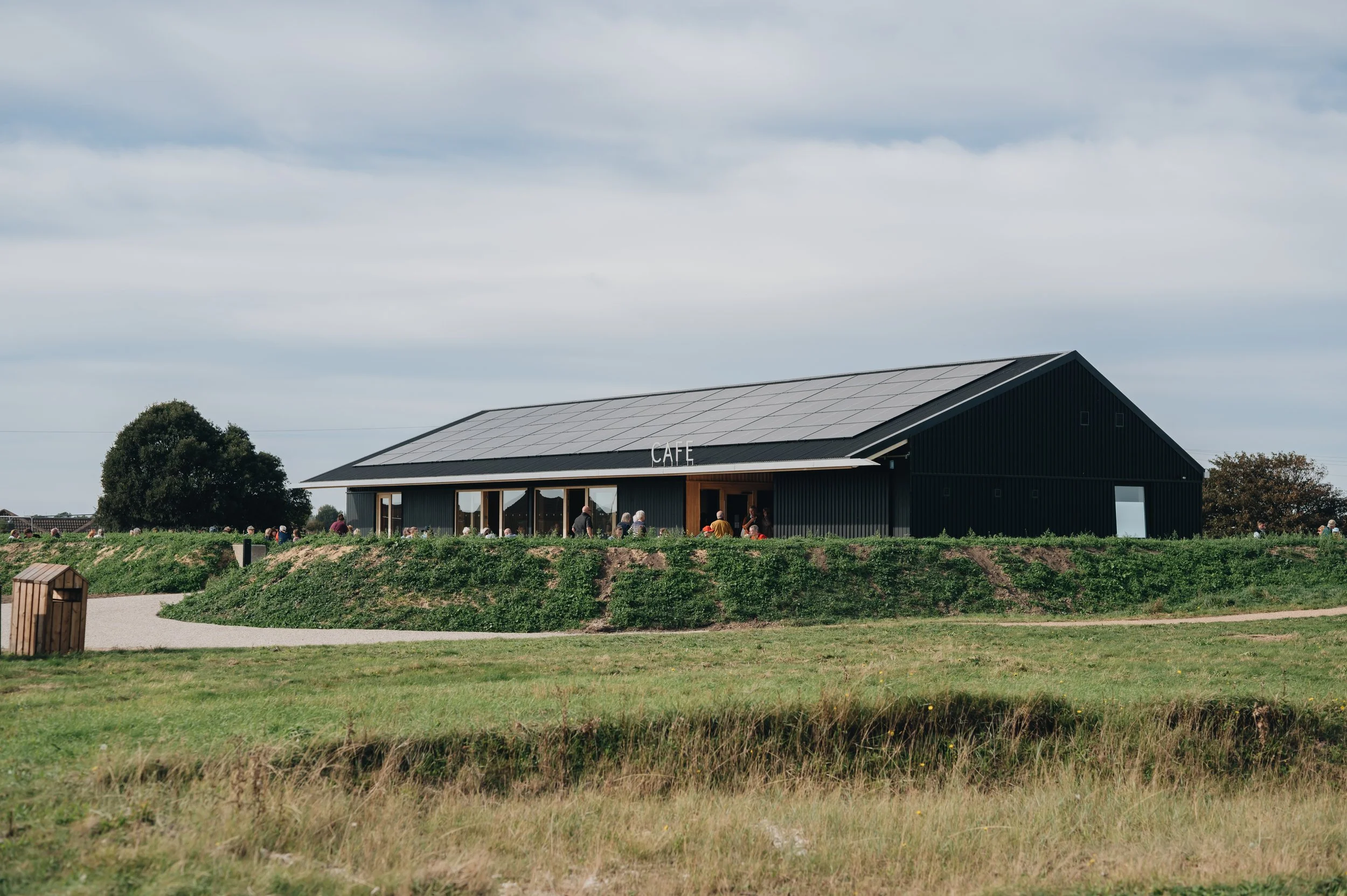 Modern black café building with solar panels on its roof, surrounded by grassy fields and people.