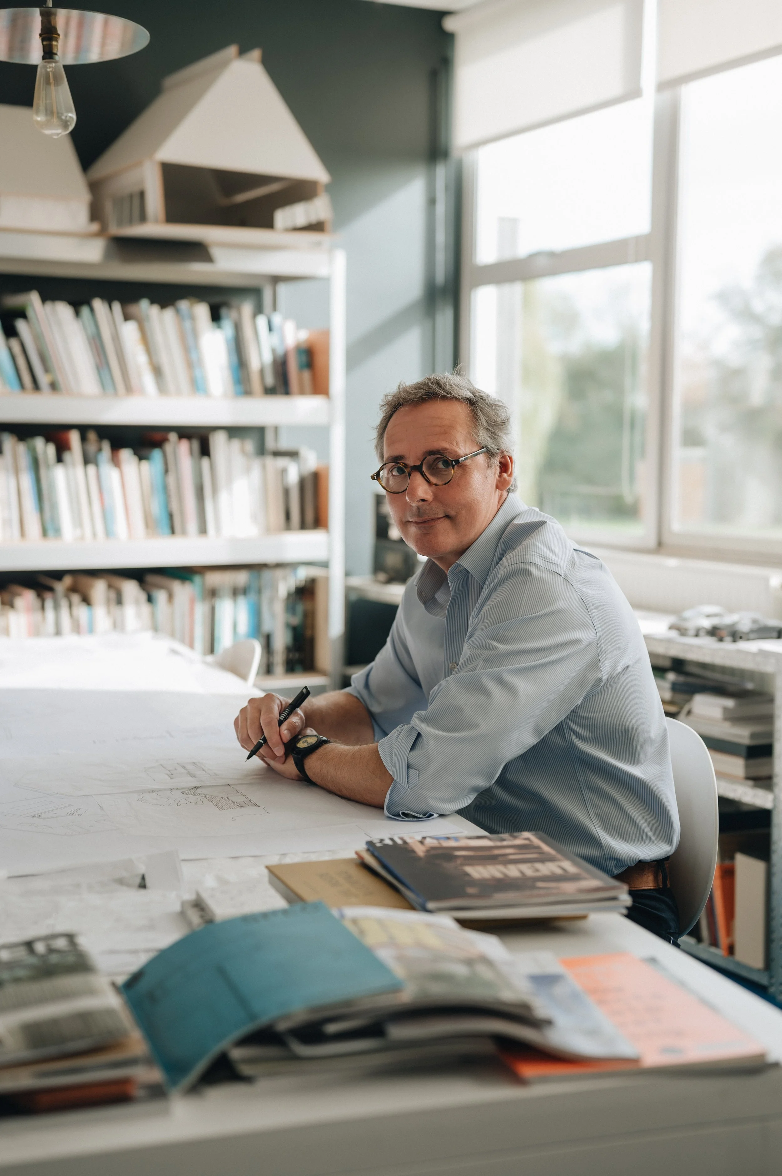 Architect wearing glasses and a striped shirt sketching at a desk in a bright office surrounded by books and architectural plans