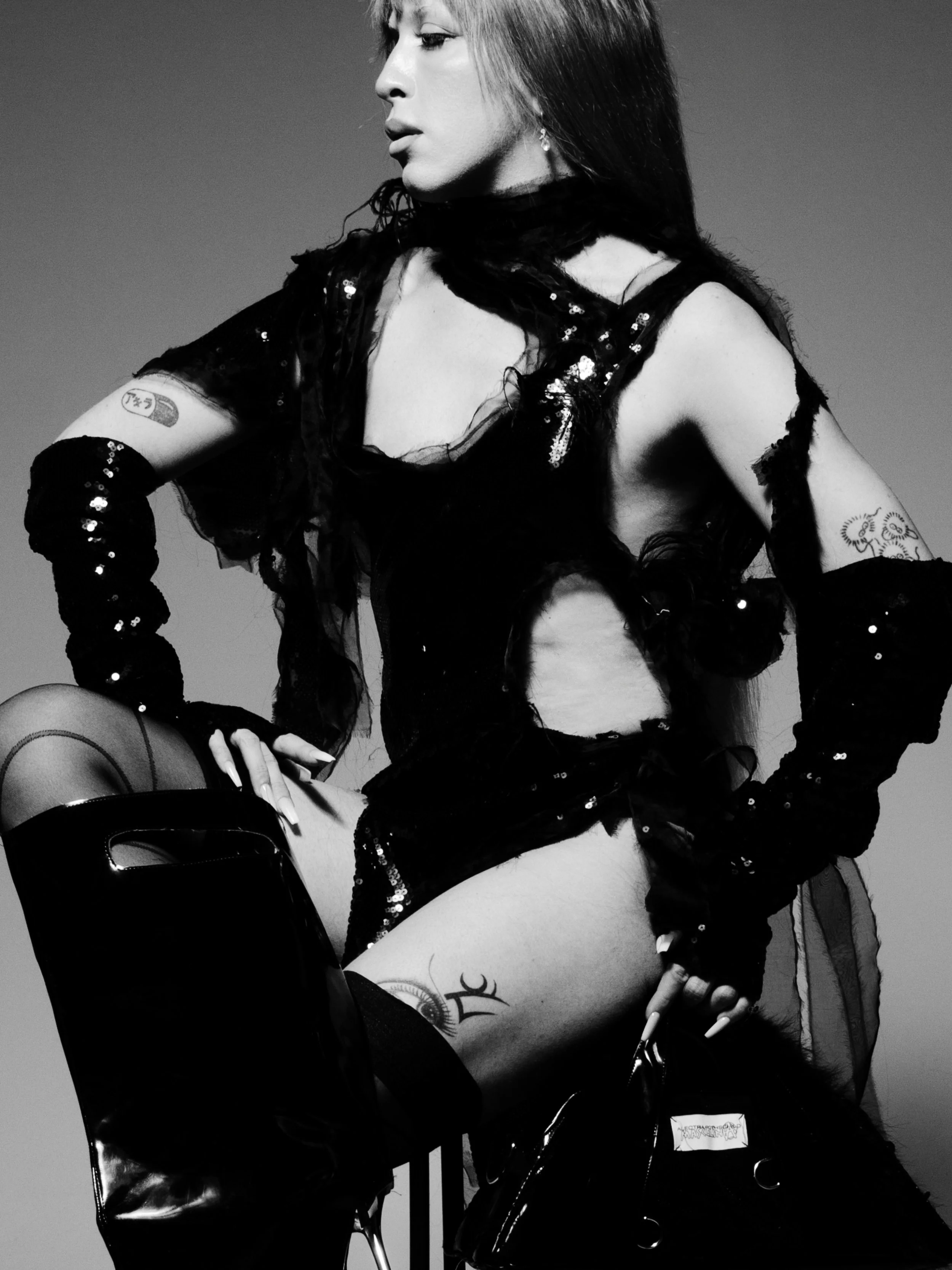 Black and white photo of a woman with long dark hair, dressed in a shiny, sequin details costume with gloves and stockings, sitting on a chair. She has tattoos on her arm and thigh, and is posed in a confident, stylish manner.