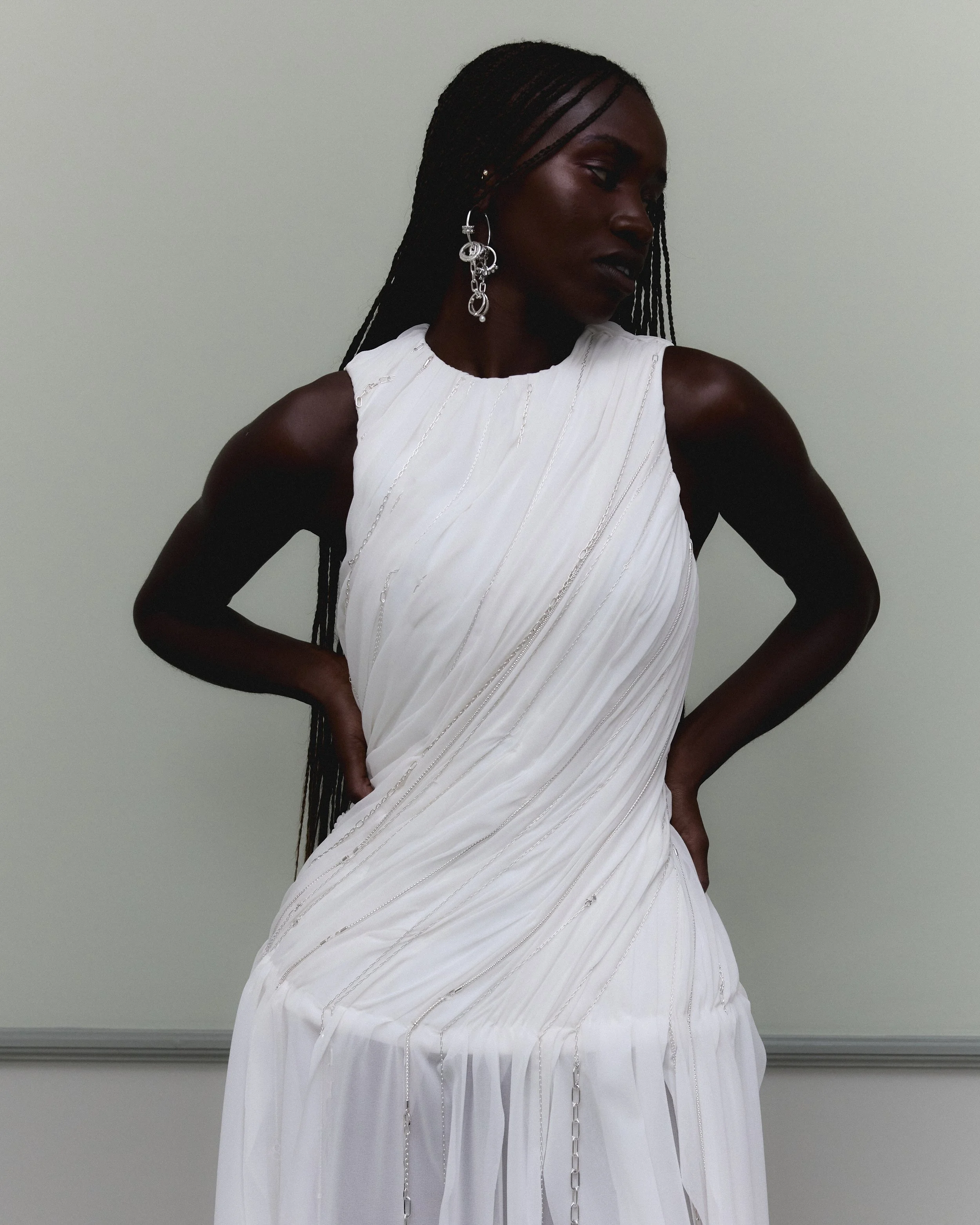 A woman with dark skin and long braided hair wearing silver jewelry, including large earrings, poses in a white dress with vertical silver thread details against a plain gray wall.