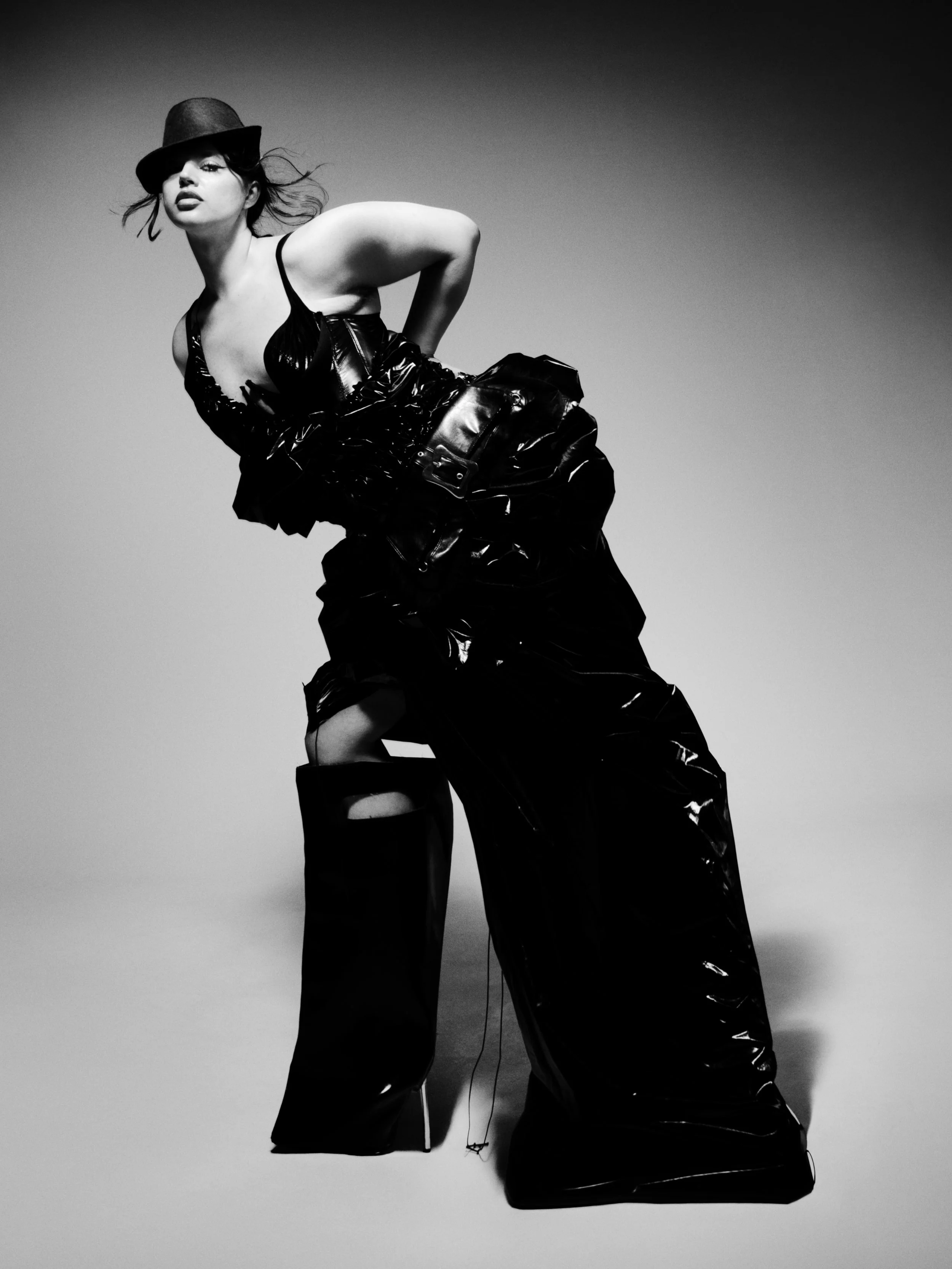 Black and white photo of a woman wearing a hat, a shiny top, and shiny pants, striking a pose against a gradient background.