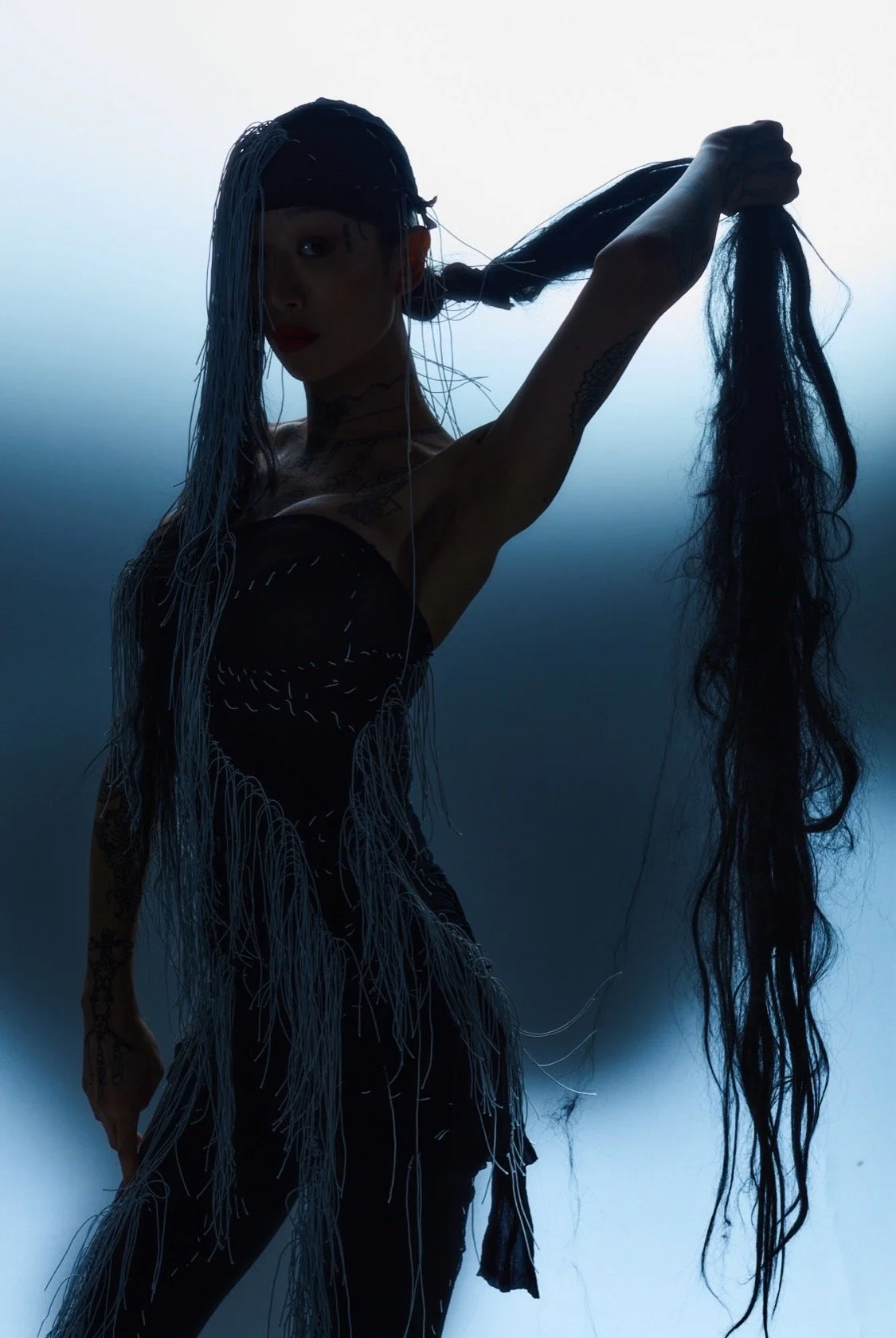 A silhouette of a woman with long black hair, held in one hand, against a blue gradient background, with visible tattoos on her arm and shoulder, wearing a black outfit with fringes.