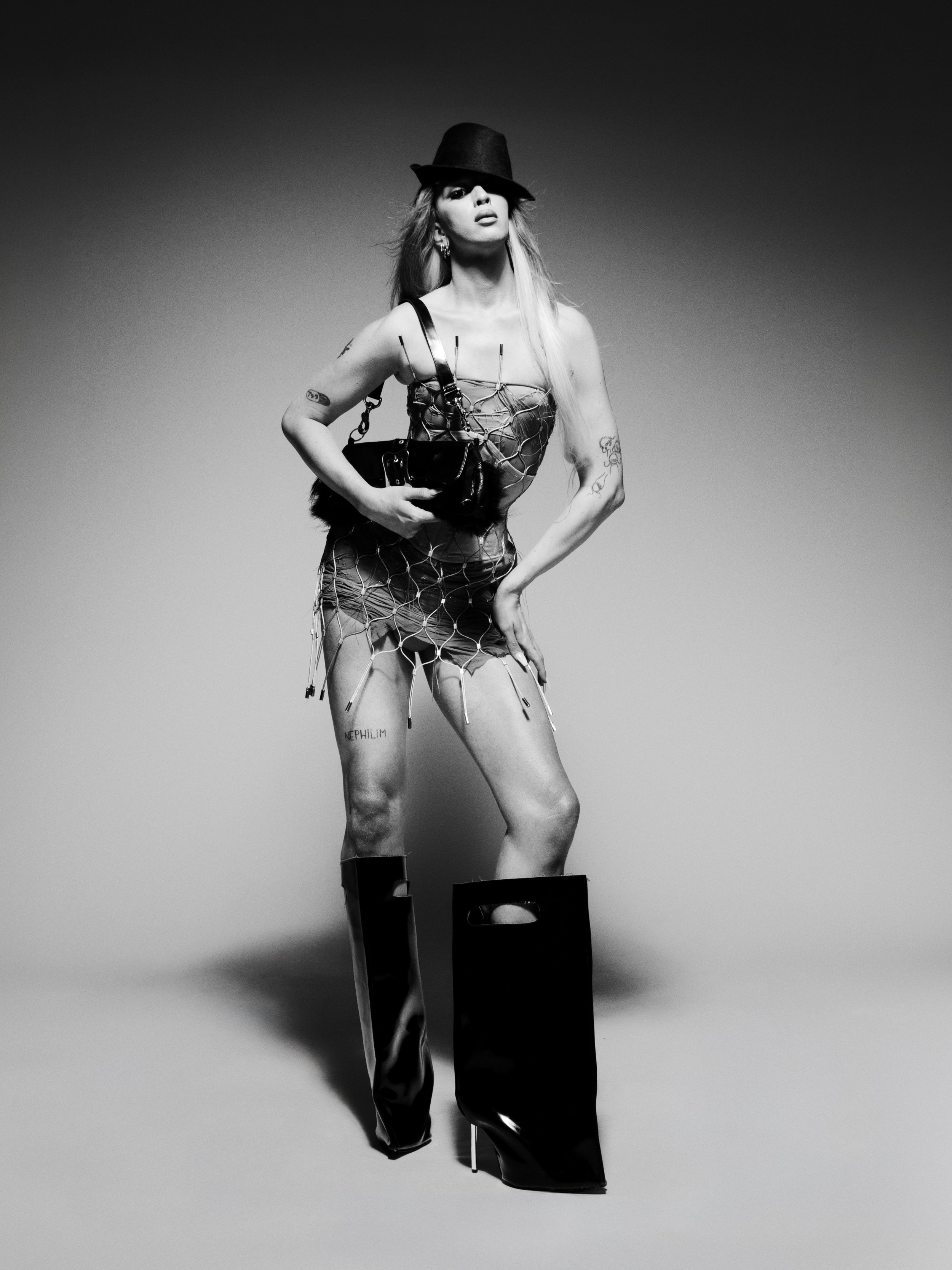 Black and white photo of a woman with long hair wearing high heels, a dress with a netted overlay, a hat covering part of her face, and holding a small bag. She has tattoos on her arms and a tattoo on her left thigh.
