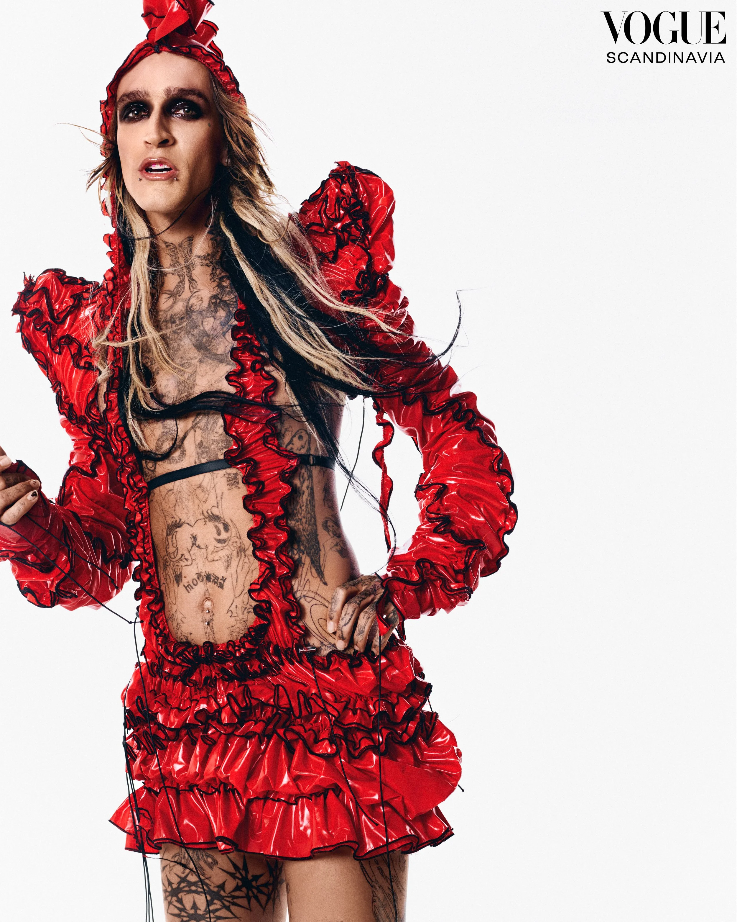 A woman with tattoos wearing a voluminous red ruffled dress and a matching headwrap, standing against a white background. She has dark makeup, a lip piercing, and is posed with her hand on her hip.