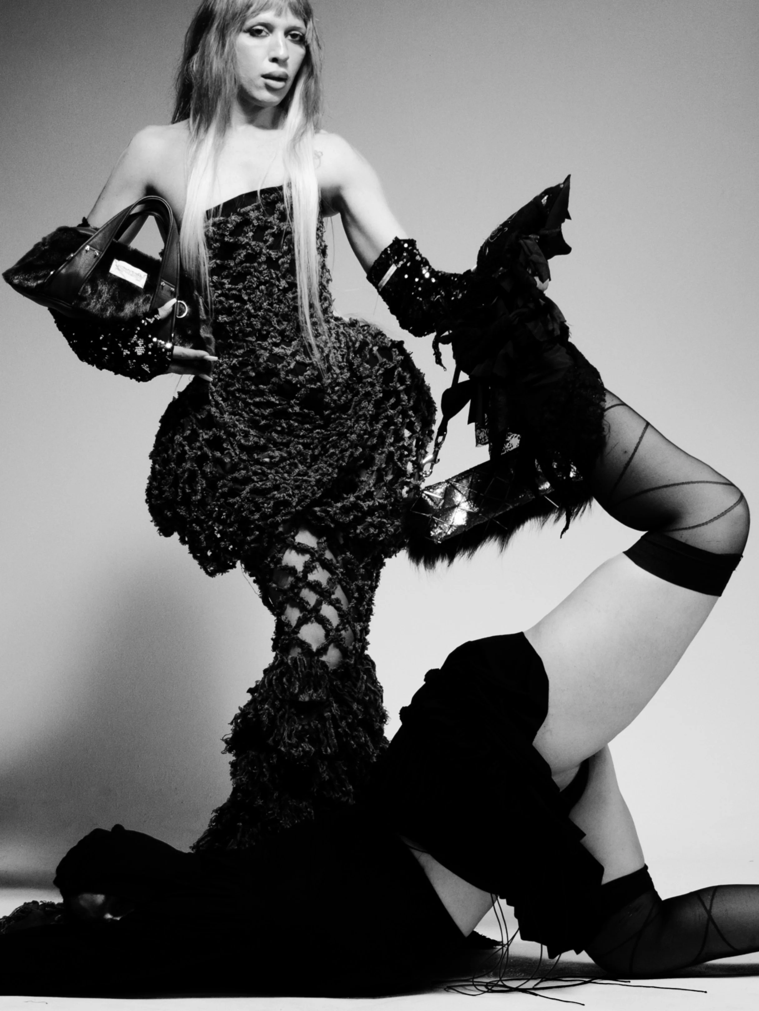 Black and white photo of a woman standing in a dynamic pose with her hands on her hips. She has long straight hair and is wearing a strapless, textured dress with sequins and lace-up shoes. There is a person lying on the floor with legs bent and wear