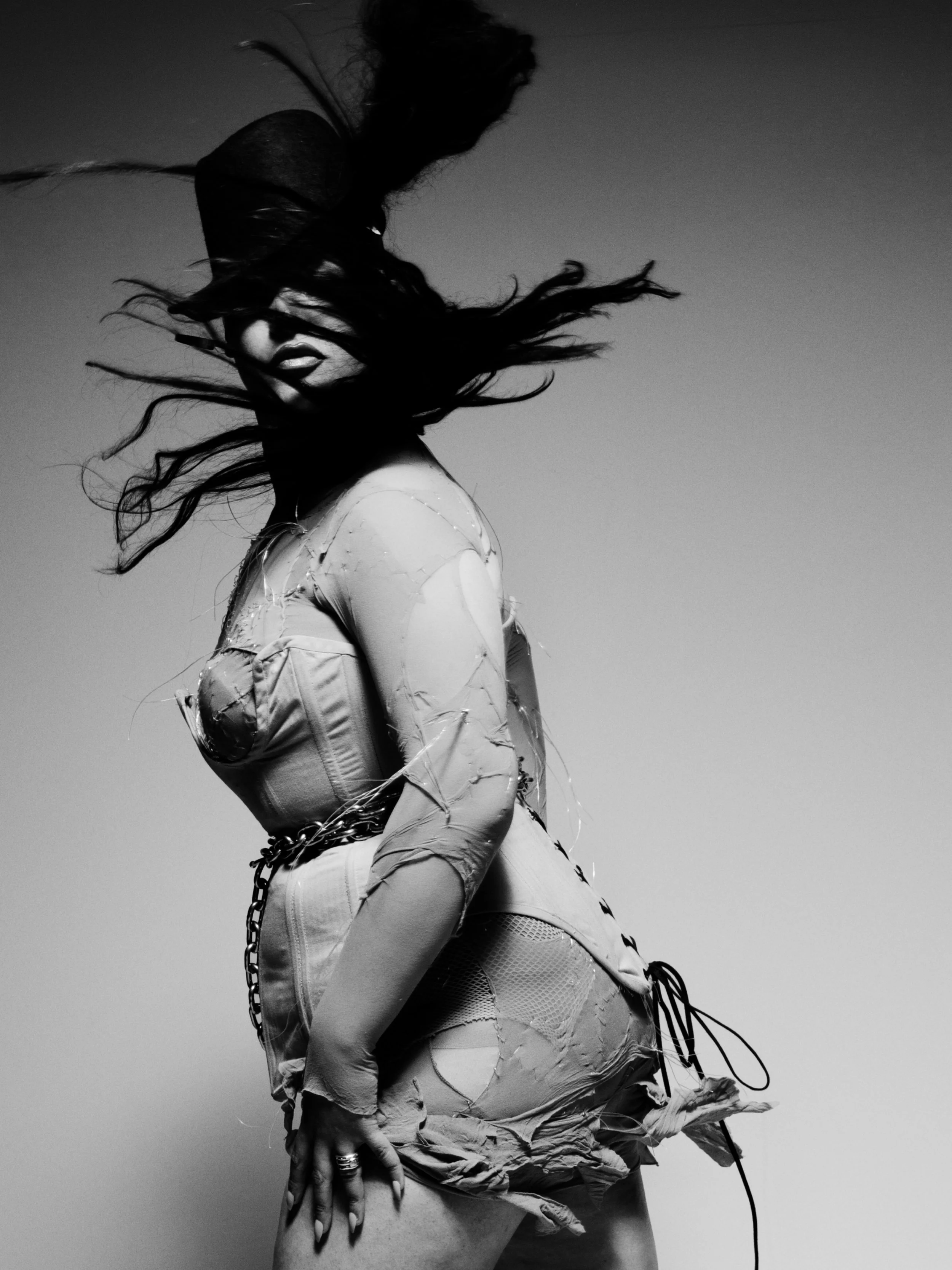 Black and white photo of a woman with long, flowing hair, wearing a ripped jacket and fishnet stockings, posed against a plain background.