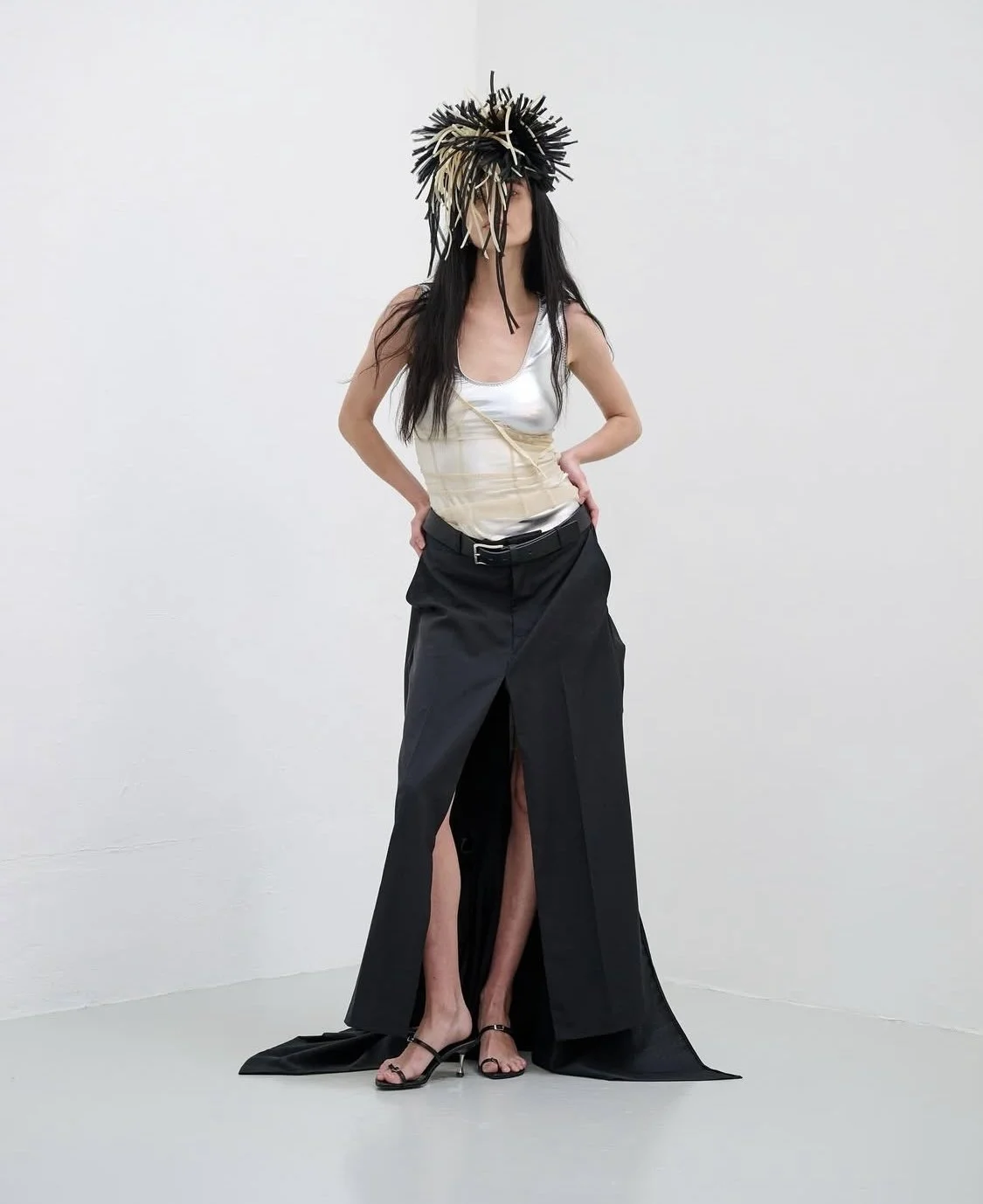 A woman with long black hair wearing a large basket-like hat made of sticks, a shiny silver top, and a long black skirt with a high slit, standing against a plain white wall.