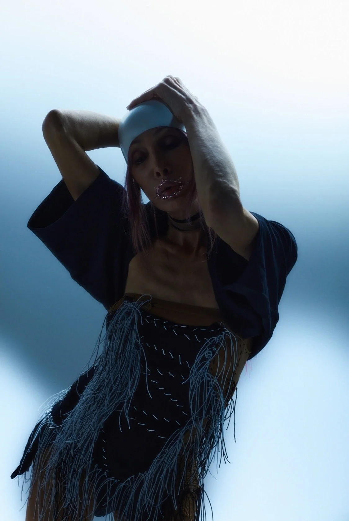 A woman wearing a white swim cap, black attire, and a fringed skirt, poses with her hand on her head against a bright blue background.