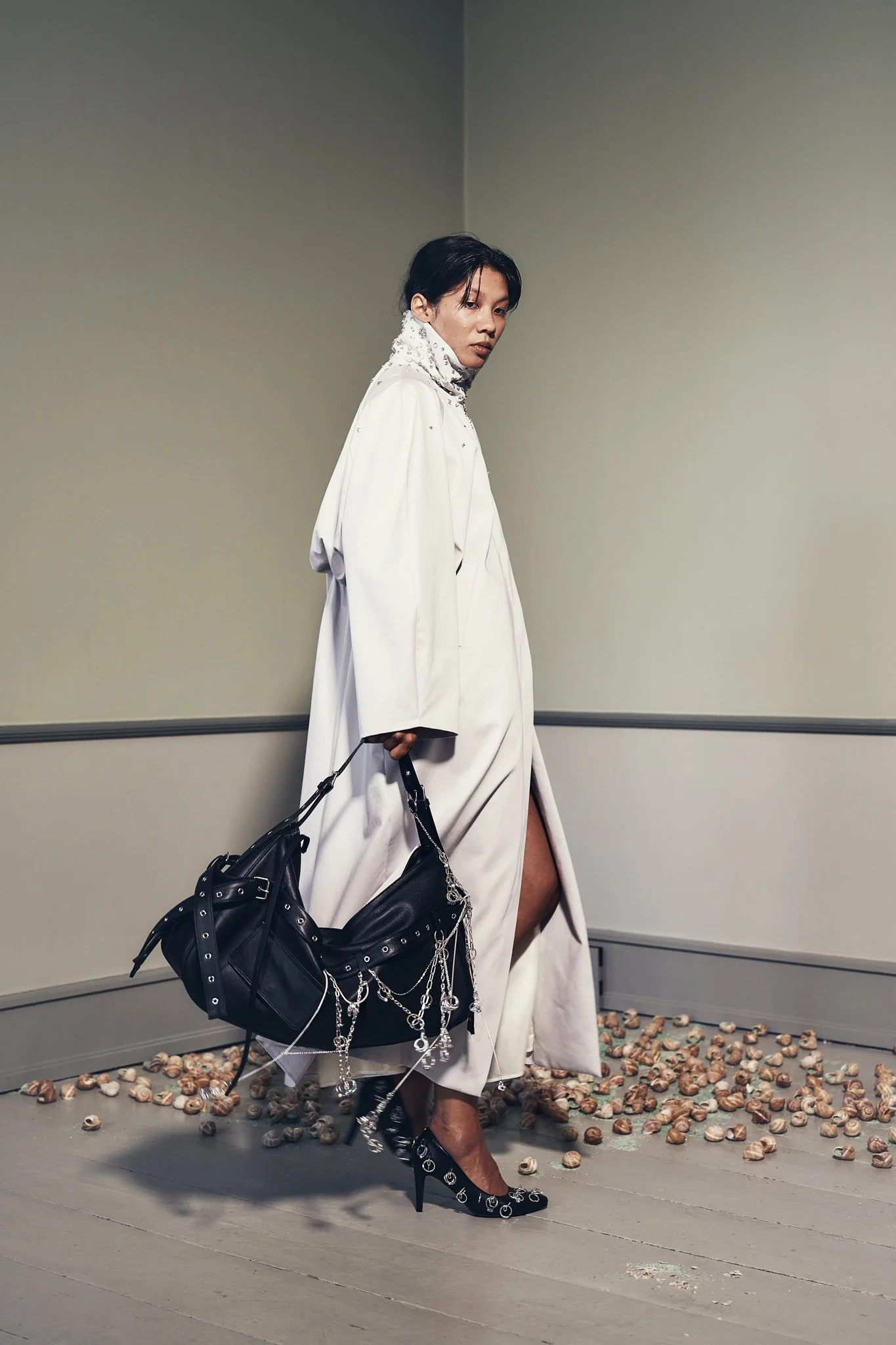 A model in a white outfit with black high heels, black handbag with silver chains, and silver jewelry, standing on a floor scattered with seashells and pebbles, against a plain pale wall.