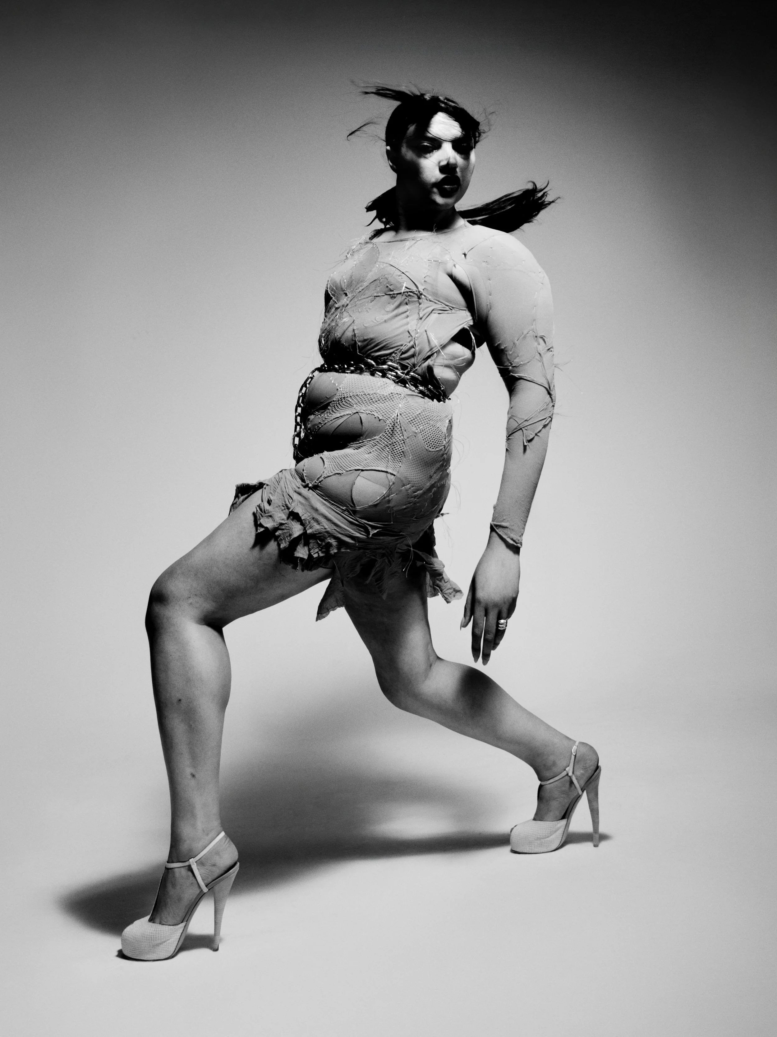 Black and white photo of a woman in high heels, wearing a ripped dress, striking a dance pose against a plain background.