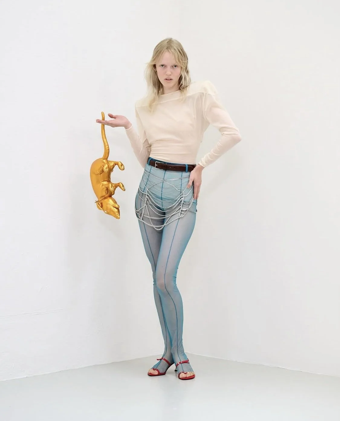 A woman with blonde hair, wearing a sheer cream-colored long-sleeve top, blue patterned tights with chain details, and red sandals, stands against a plain white wall. She holds a gold inflatable sculpture resembling an animal.