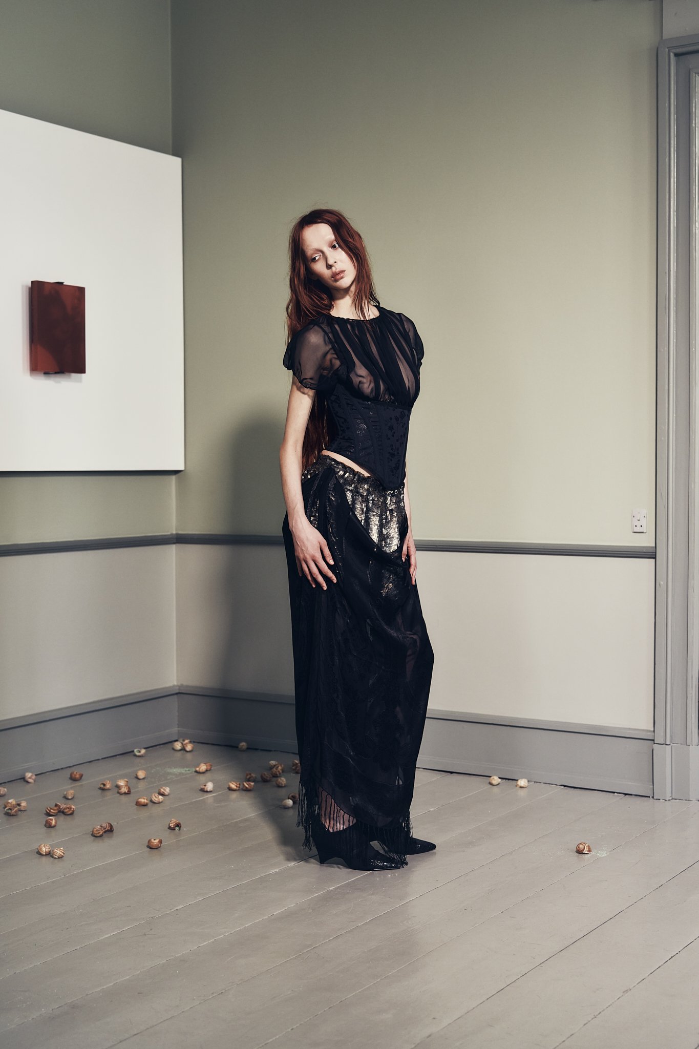 A woman with long red hair wearing a black sheer top and long black skirt stands in a room with scattered nuts on the floor, beige walls, a white panel, and a brown wall decor piece.