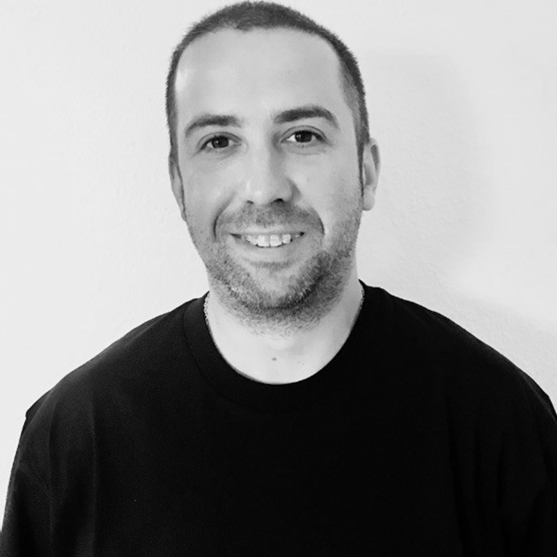 Black and white portrait of a smiling man with short hair, wearing a black t-shirt, standing against a plain wall.