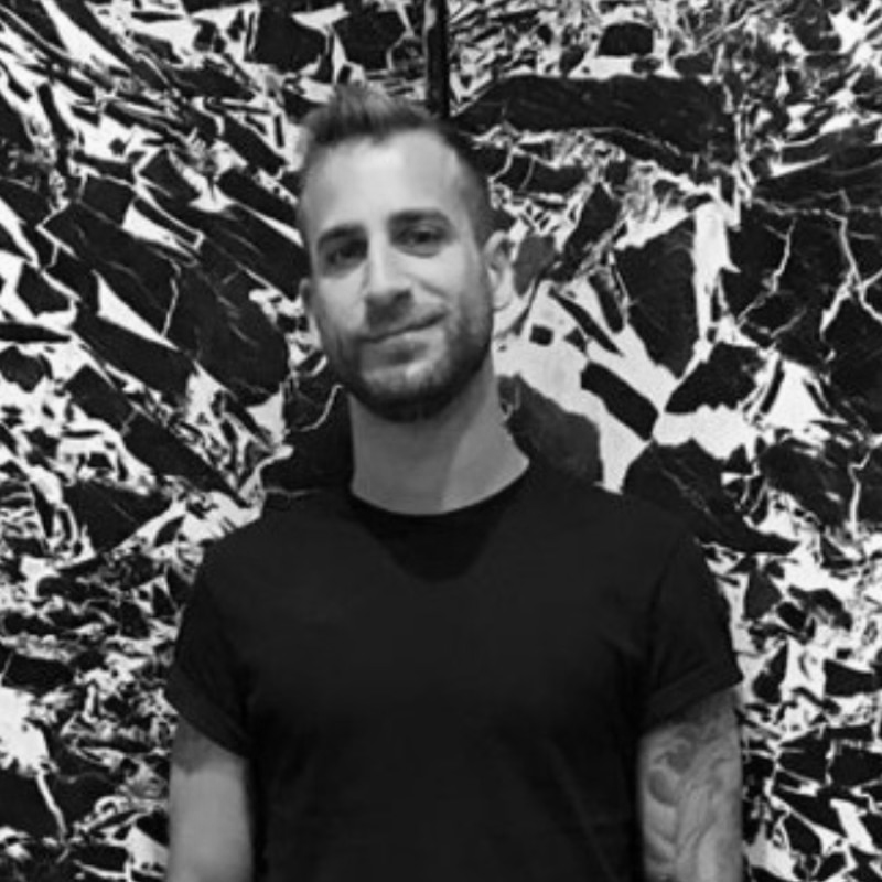 Black and white photo of a man with short hair and a beard, wearing a black t-shirt, standing in front of an abstract, artistic background that resembles a highly textured, monochrome forest or jungle scene.