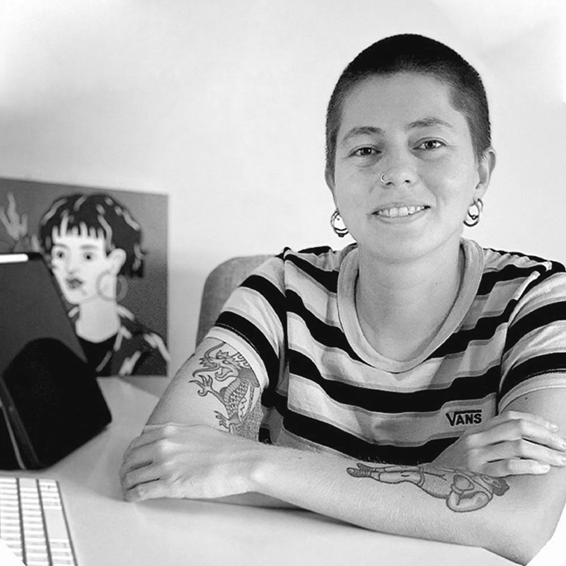 A person with a buzz cut hairstyle, wearing a striped t-shirt and earrings, sits at a table with arms crossed, smiling at the camera. They have tattoos on their arms, including a dragon and a shell. In the background, there is a framed art piece of a stylized female face.