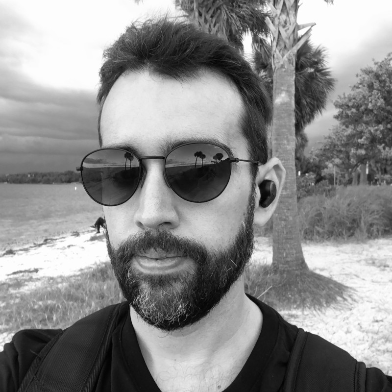 A black-and-white photo of a man with sunglasses and a beard taking a selfie outdoors near a beach, with palm trees and water in the background.