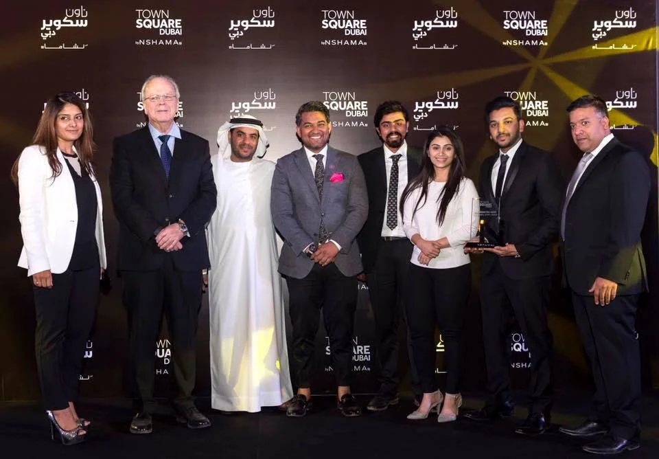 Group of eight people standing together at an event with a backdrop displaying 'Town Square Dubai' and 'Nshama'. They are dressed in formal attire, with some holding an award or trophy. The group appears to be celebrating or recognized for an achievement.