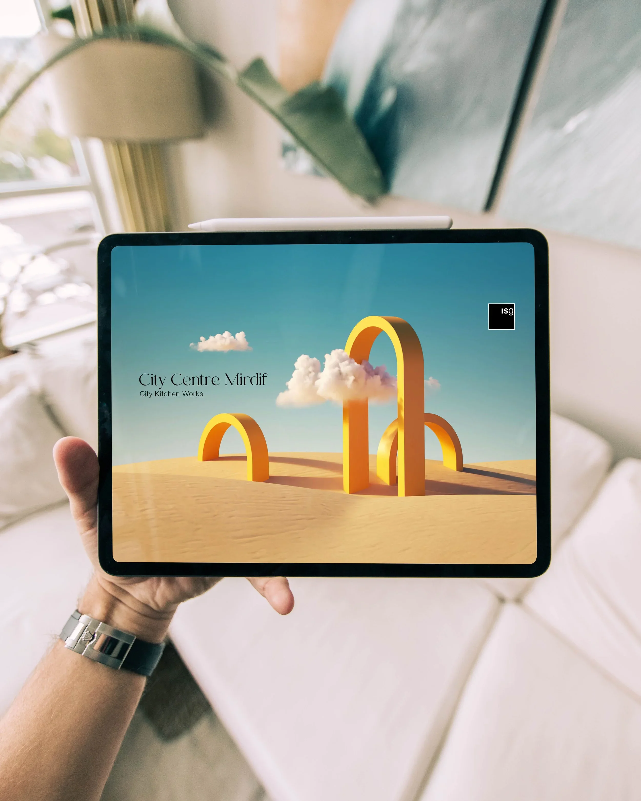 Person holding a tablet displaying colorful abstract graphic titled 'City Centre Mirdif' with a cloud and rainbow arch, indoors near a window.