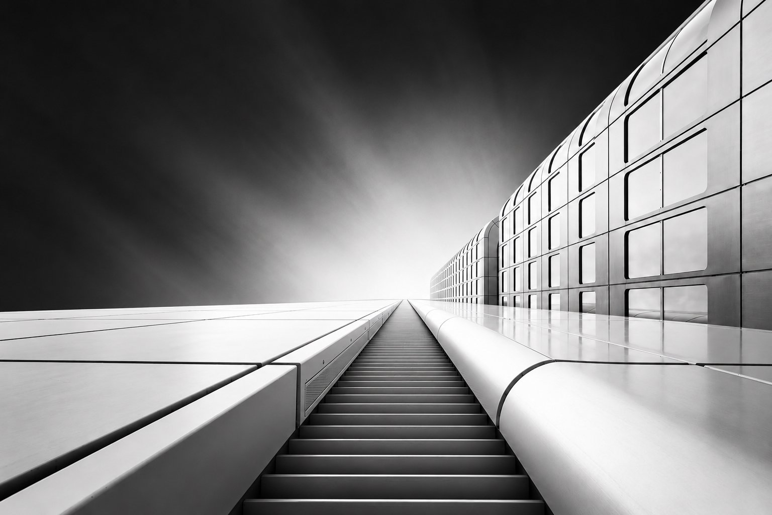 Black-and-white architectural photograph of a modern skyscraper with a staircase rising toward the sky, emphasizing geometric lines and perspective.