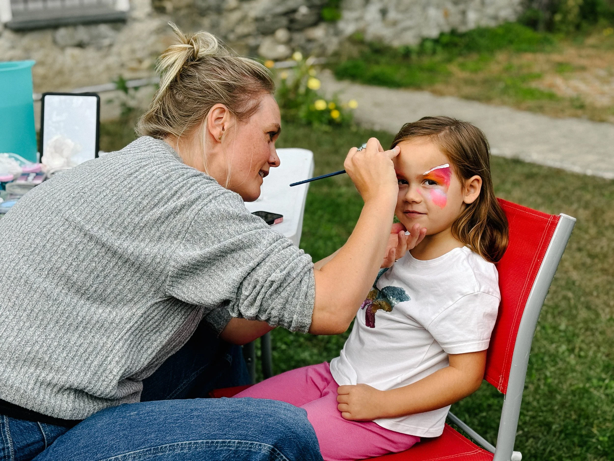 A woman with blonde hair styled in a high messy bun is painting a young girl's face with bright colors, sitting outdoors on a red chair. The girl has a face painting design in pink, orange, and white. The background shows a garden with a stone wall and yellow flowers.