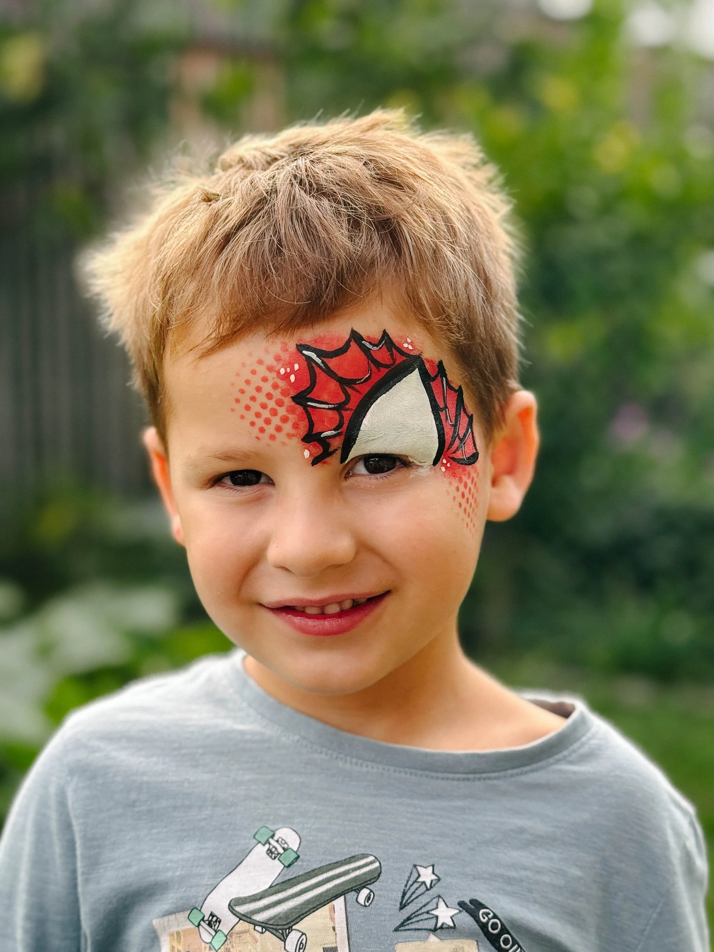 A young boy with painted face featuring a superhero mask design. The face paint includes a white mask outlined in black, with red and black web patterns around the eyes. The boy has light brown, tousled hair and is wearing a gray T-shirt with skateboarding-themed illustrations.