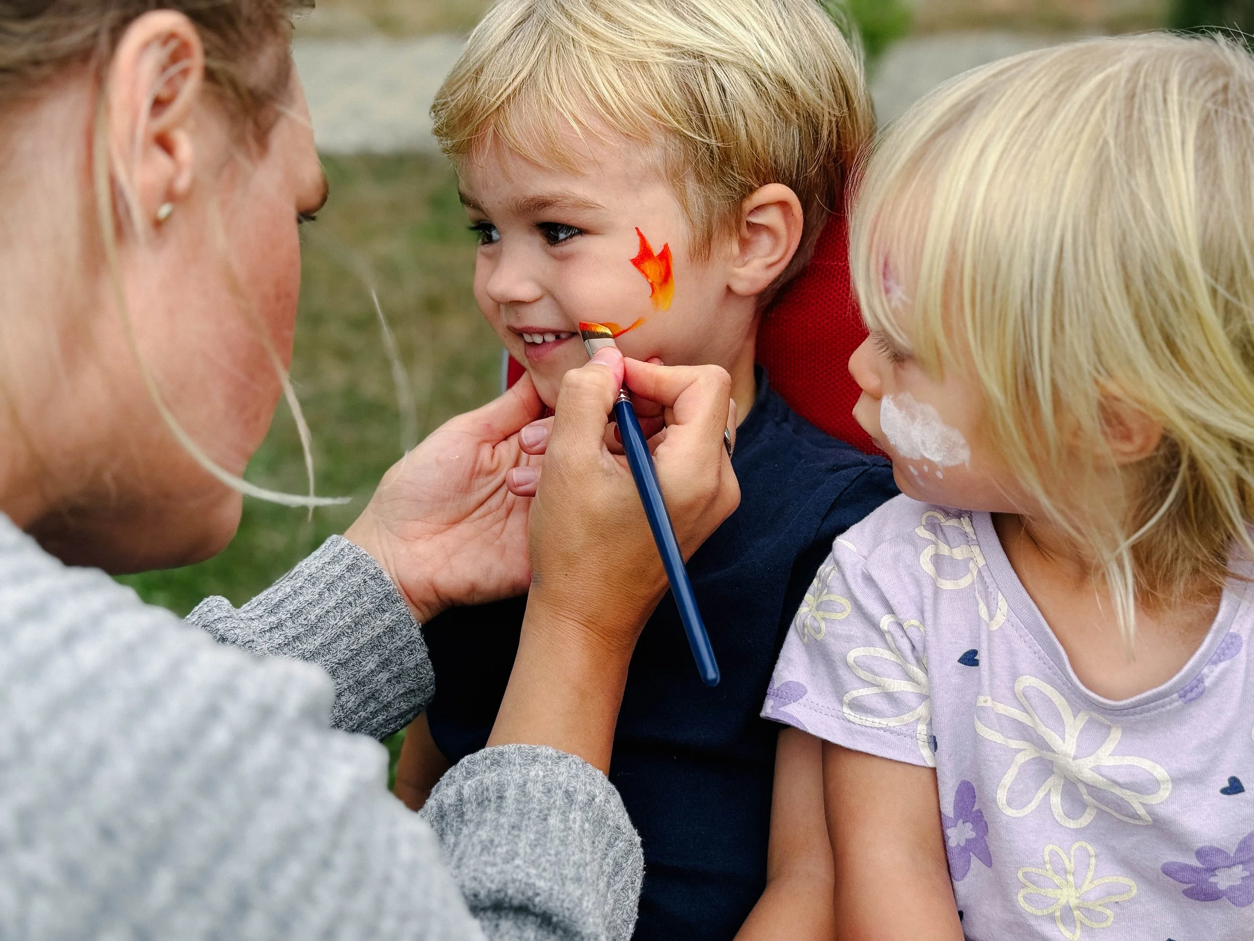 A woman is painting a fire on a young boy's face with face paint, while another girl with blonde hair looks on.