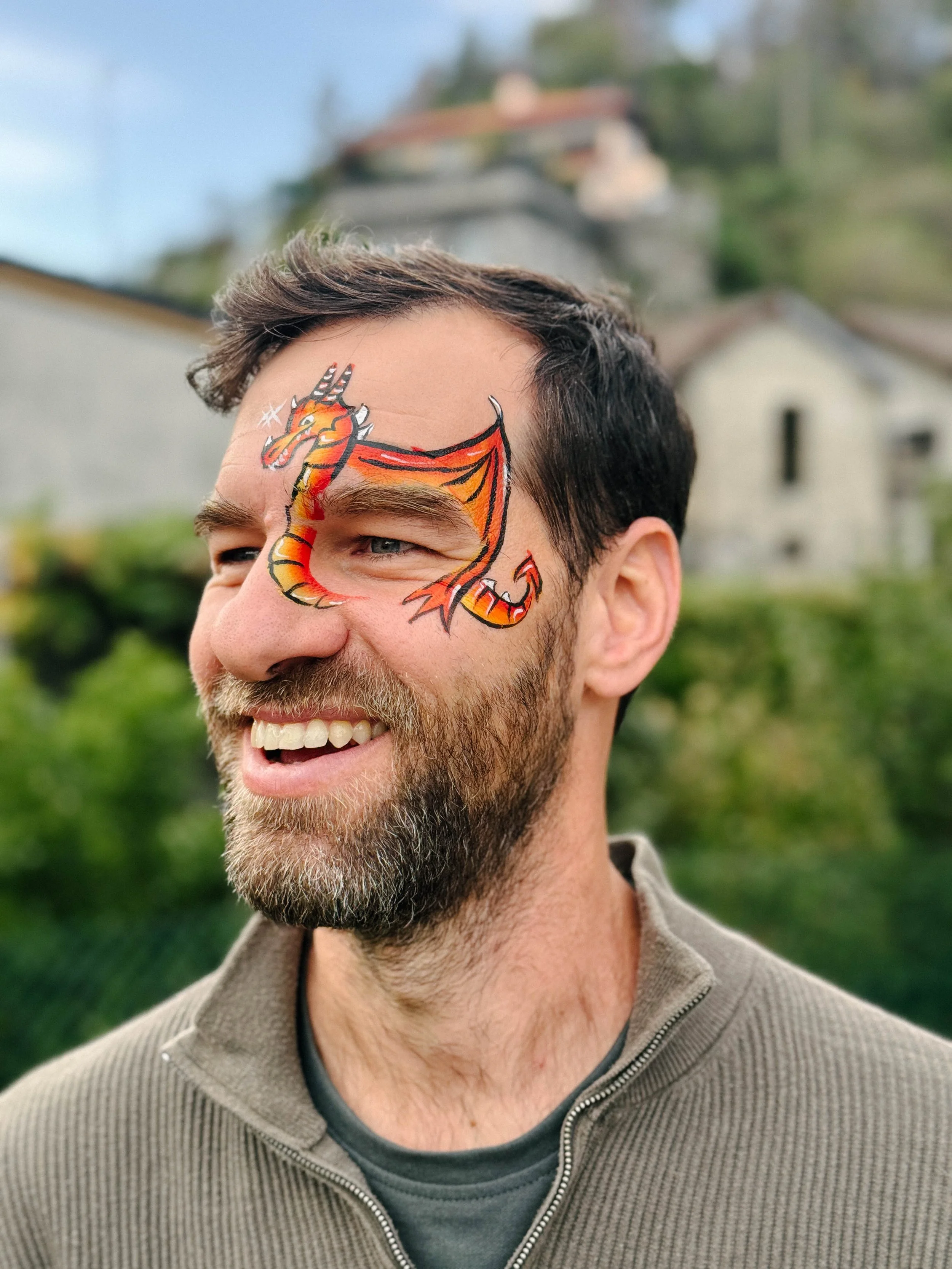 Close-up of a smiling man with a dragon face face paint on his face, outdoor background with trees and cloudy sky.
