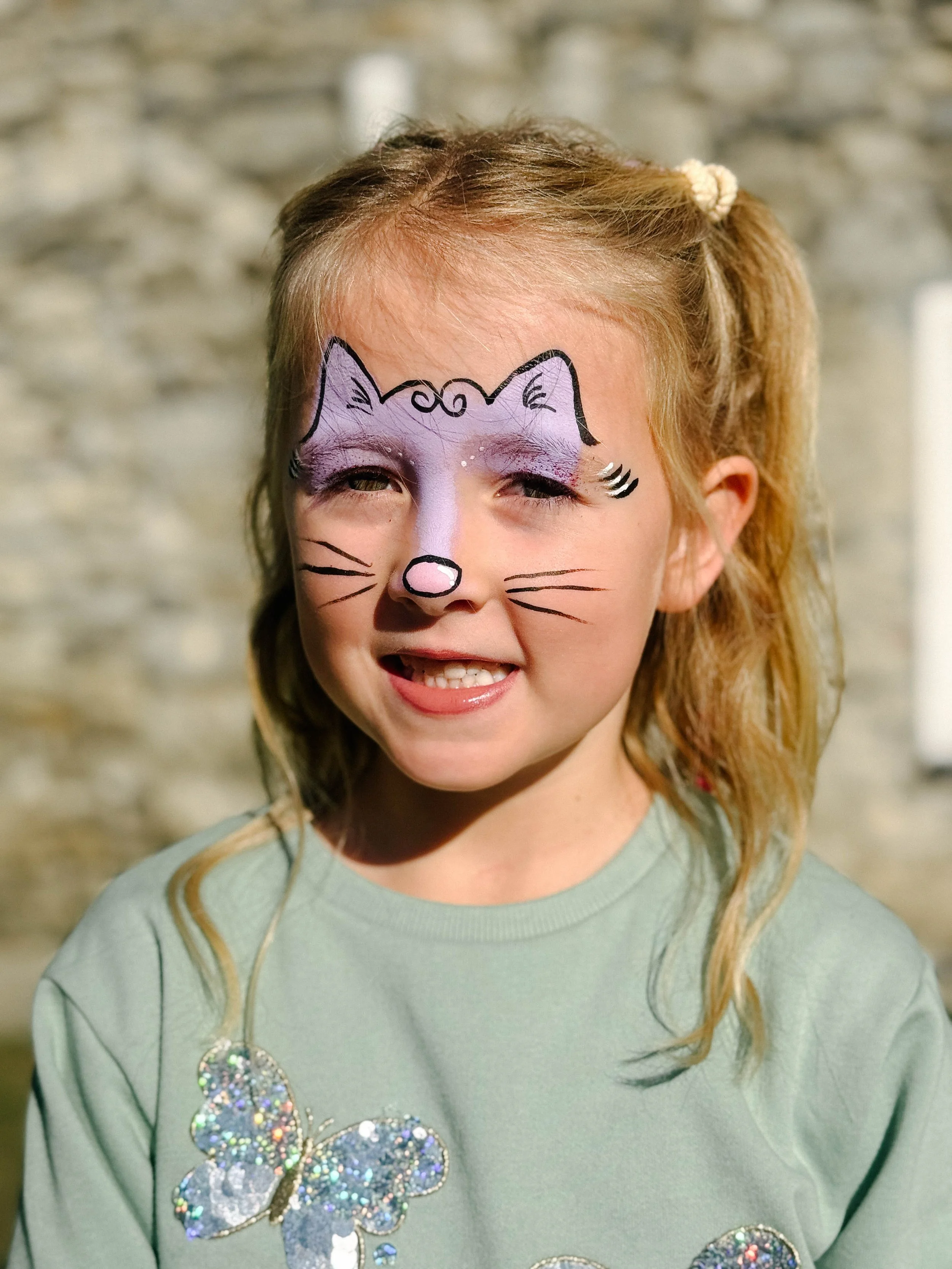 A young girl with face paint resembling a purple and pink cat with black outlines and whiskers. She is smiling and has light brown hair tied in a ponytail with a cream scrunchie, wearing a light green shirt with colorful butterfly designs.