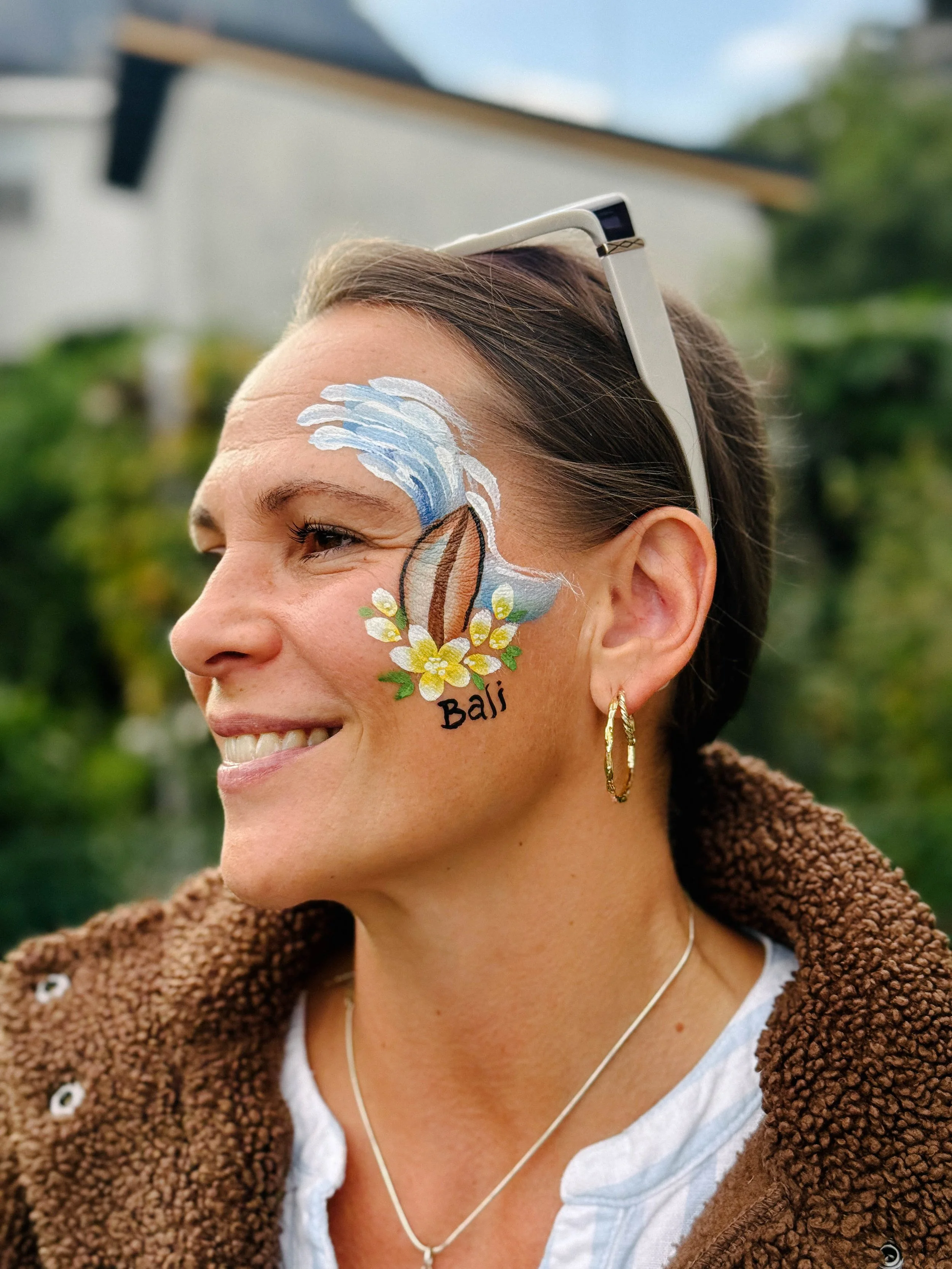 Close-up of a woman with painted face art of a wave, flowers, and the word 'Bali'. She is smiling, wearing hoop earrings, a necklace, a white t-shirt, and a brown fleece jacket, outdoors with blurred green trees in the background.