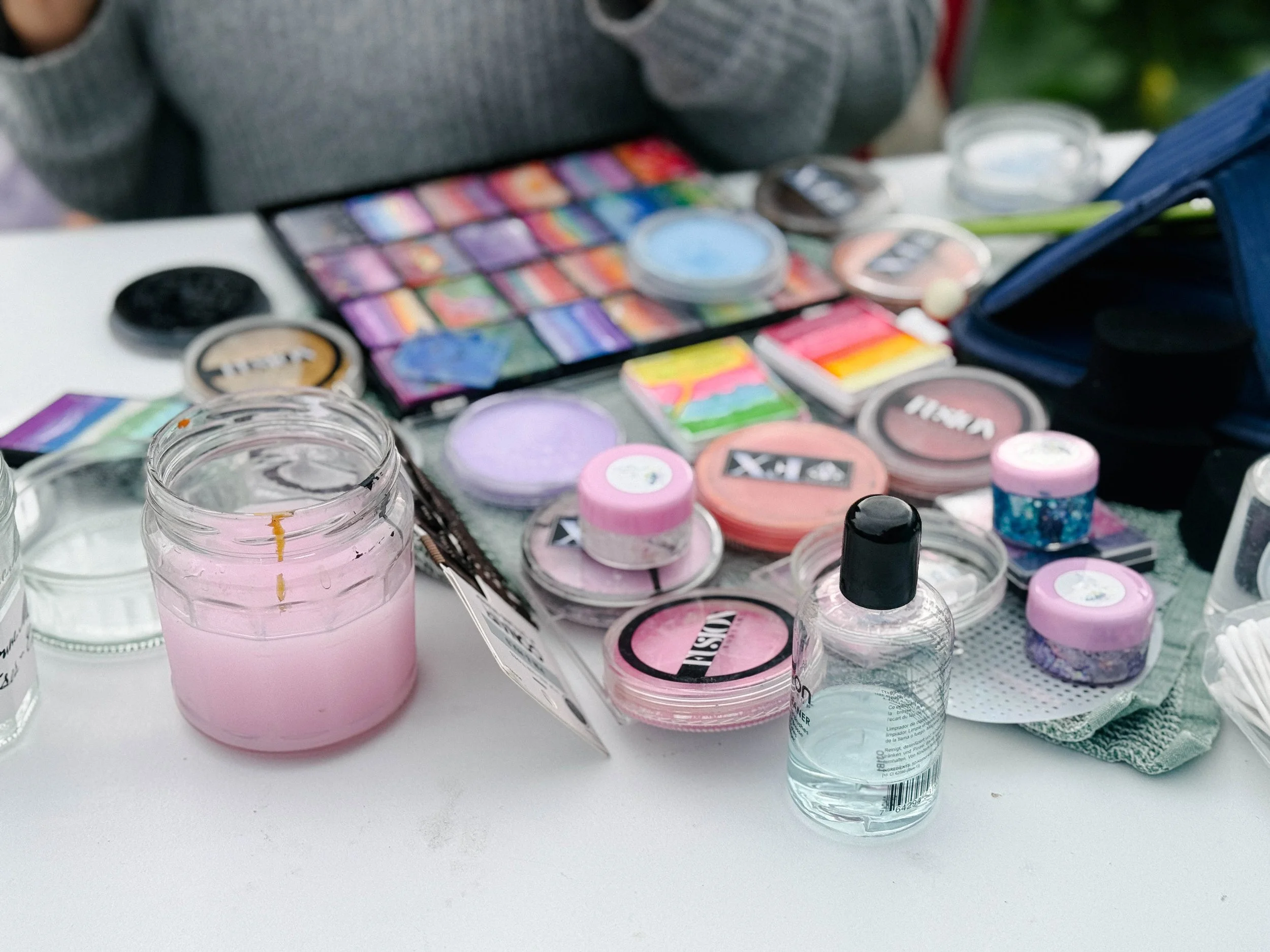 Assorted makeup products, including eyeshadows, blushes, and glosses, arranged on a white surface with some open jars of pink cream and bottles of transparent liquid.