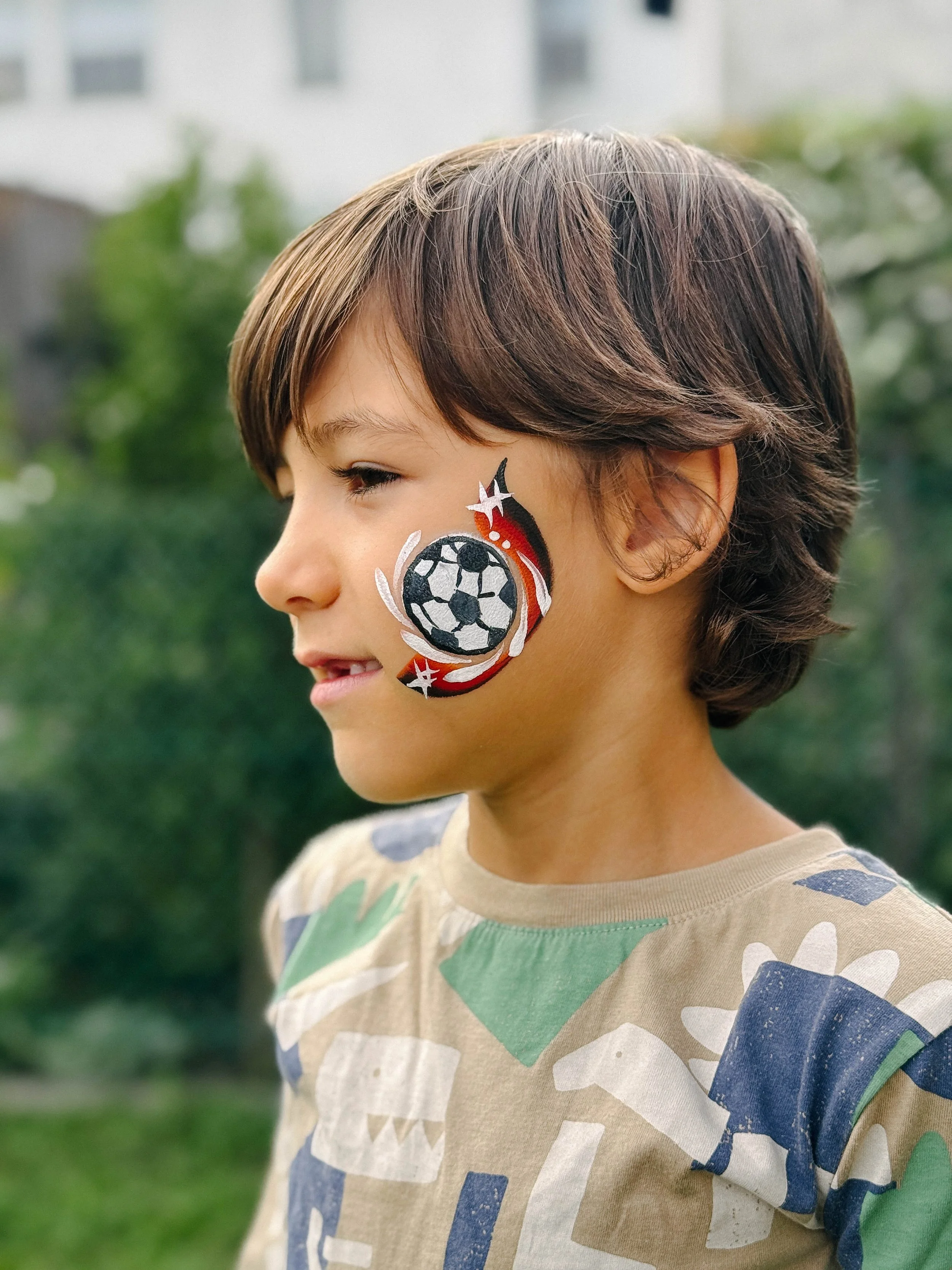 A boy with medium-length brown hair with highlights, wearing a beige T-shirt with a colorful abstract pattern, has a football-themed face painting on his right cheek, featuring a soccer ball and red and white flames.