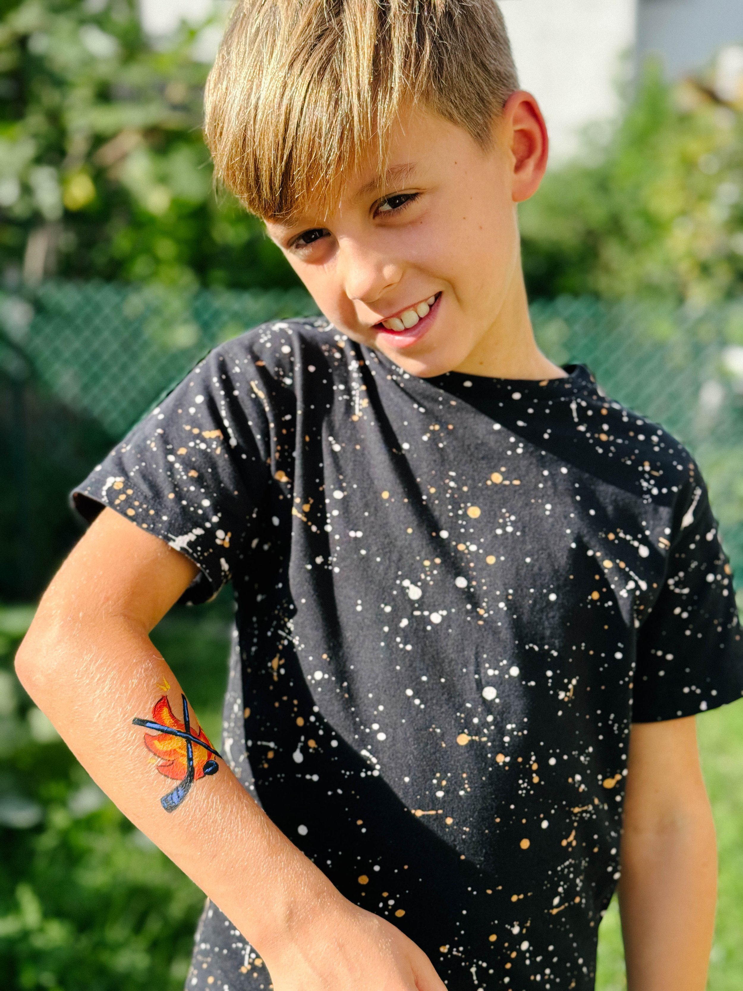 Young boy with short light brown hair, smiling, wearing a black t-shirt with white and gold speckle pattern, standing outdoors with green foliage background. He has a colorful butterfly tattoo on his left arm.