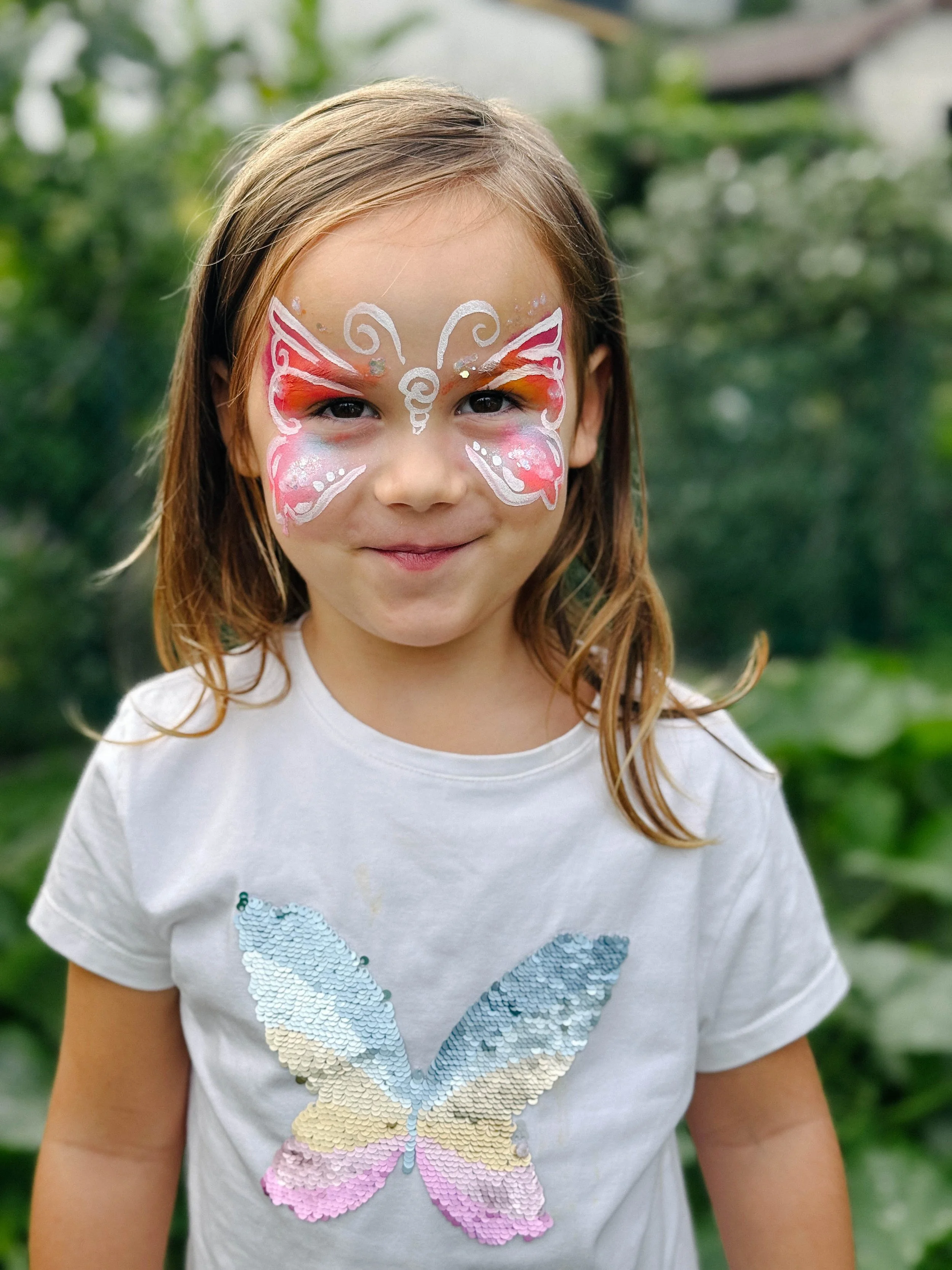 A young girl with face paint resembling a butterfly, standing outdoors with greenery in the background, wearing a white T-shirt with a sequin butterfly design.