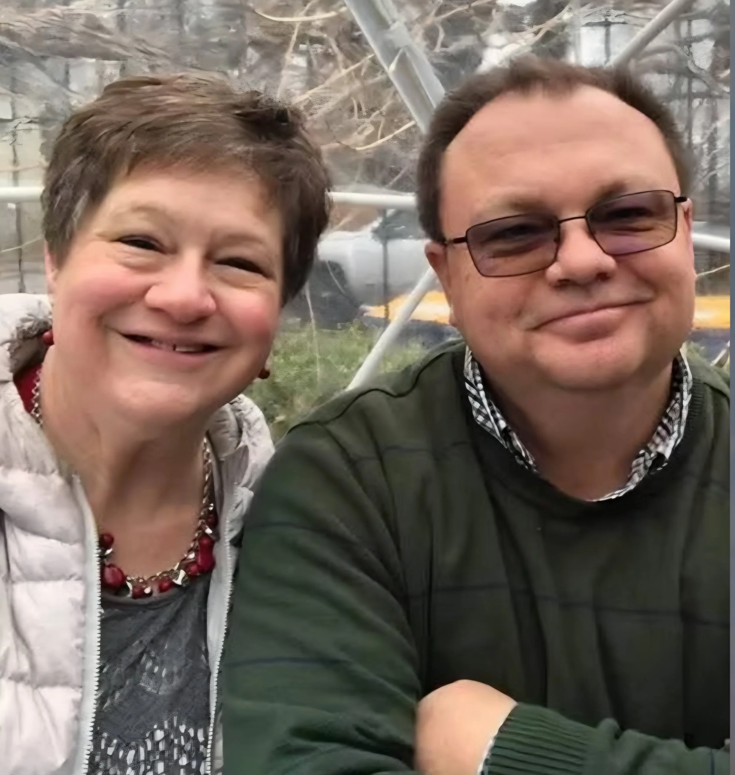 Smiling couple taking a close-up selfie indoors, with one person wearing sunglasses and a red beaded necklace.
