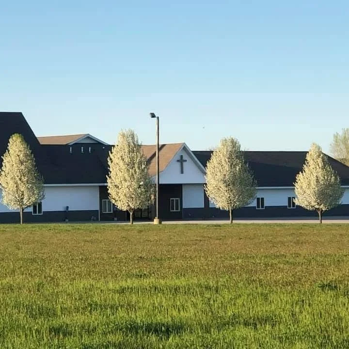 White and dark gray church with a cross, trees, lamppost, and grassy lawn under a clear blue sky
