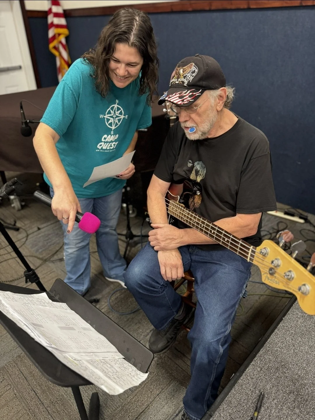 Woman holding a pink microphone beside an older man playing bass guitar in a rehearsal room.