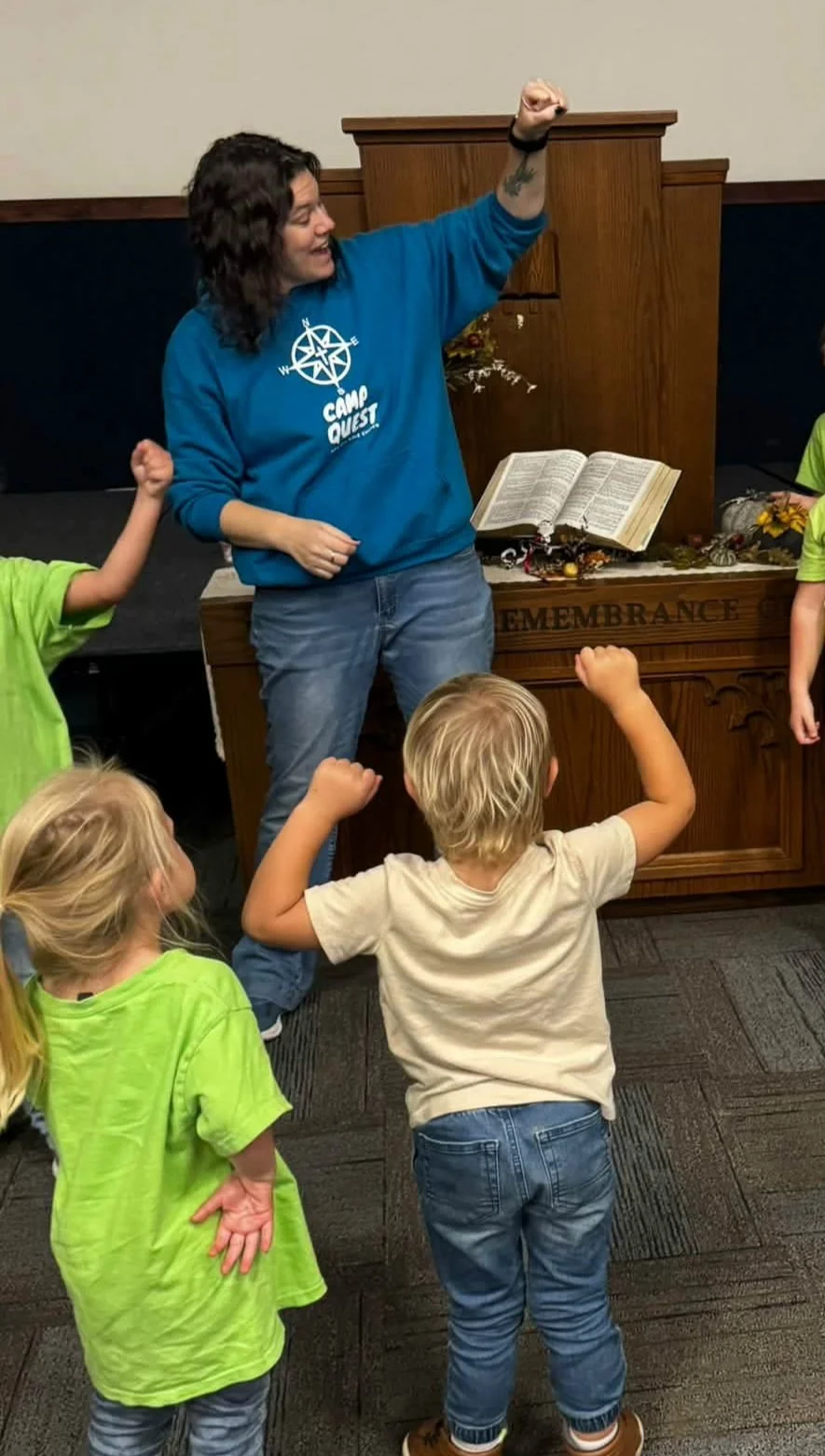 Children raise their hands during a camp activity led by a woman in front of a church pulpit with an open Bible.